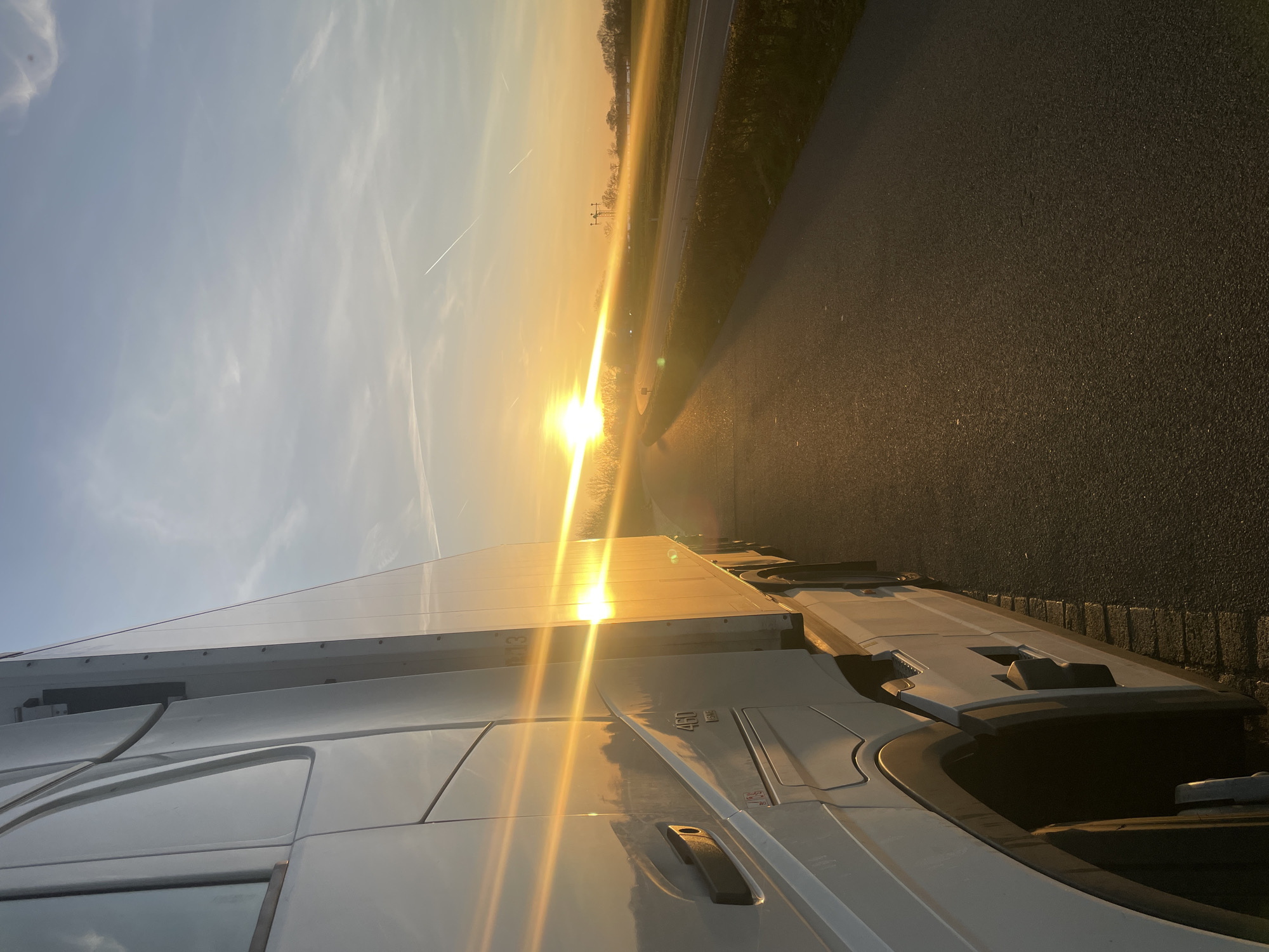 The image shows a sunlit scene in Volkel, Netherlands, featuring a truck parked on the roadside. The sun is low on the horizon, casting a bright glare. A road and a grassy area are visible beyond the truck. There are no people in the frame. The sky is partially clear with a few clouds and contrails.