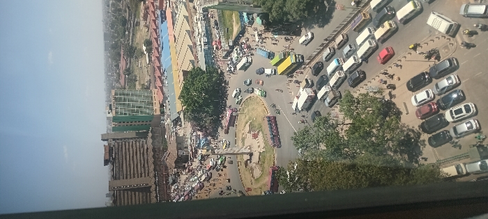 The image depicts a high-angle aerial view of a densely populated urban intersection in Nairobi, Kenya, under a clear, bright daytime sky. The central feature is a large, circular multi-lane roundabout with a tall, light-colored, tapering monument situated in its grassy core. This roundabout is identified as the Uhuru Highway / Haile Selassie Avenue Roundabout, located at the south-western edge of Nairobi's Central Business District. The perspective is oriented generally towards the north-northeast.
Heavy vehicular traffic, including numerous cars (sedans, SUVs), white and blue "matatu" minibuses, yellow and green large buses, and a few motorcycles, occupies all lanes of the roundabout and the radiating feeder roads. Vehicles are observed in various states of motion and queuing.
Pedestrians are visible on sidewalks and attempting to cross streets surrounding the roundabout, particularly on the right side of the image.
In the mid-ground, beyond the roundabout, a sprawling complex of low-rise structures with multi-colored corrugated iron roofs (blue, red, yellow) is prominent, consistent with a market area such as the Globe Roundabout Market or Gikomba Market.
The background features several multi-story buildings characteristic of an urban environment. A prominent tall, brown building with a textured facade is visible to the left of the market complex, alongside a modern, multi-story green building with a geometric pattern on its facade. Further urban development extends into the hazy distance.
In the foreground, a tree-lined street includes a dedicated parking area with numerous parked private vehicles. The left edge of the image is obscured by a window frame or structural element, indicating the photo was taken from within a building.