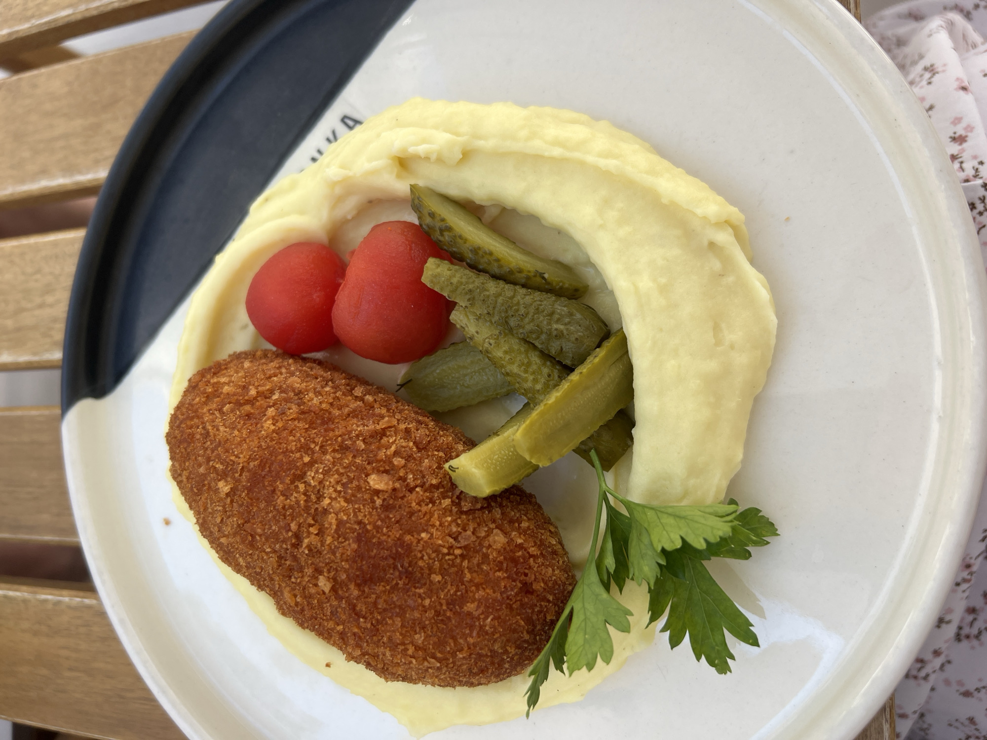 A top-down shot shows a white plate with a dark blue rim, arranged with food on a wooden table, possibly outdoors. The plate contains a breaded cutlet (likely chicken or pork), a mound of mashed potatoes encircling a few pickled tomatoes and sliced pickled cucumbers. A sprig of parsley garnishes the plate. A floral-patterned cloth is visible at the edge of the frame. While the image lacks contextual clues to verify the location, it is hypothesized to be from Boreti, Montenegro, based on the request.