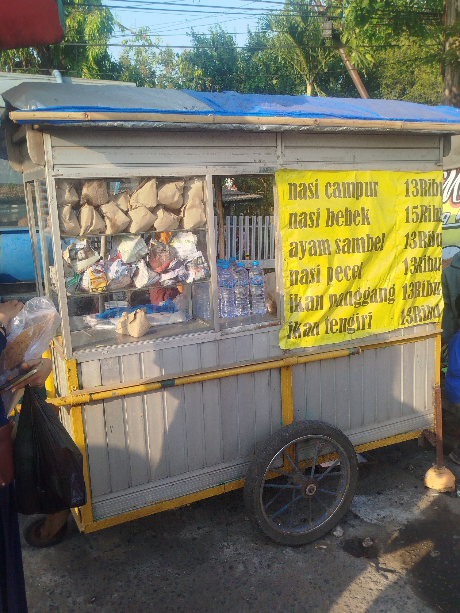 A roadside food cart is parked on a paved surface, likely a street or sidewalk, in Burneh, Indonesia. The cart is constructed from metal and wood, with a blue tarp covering the roof. A yellow banner displays a menu in Indonesian, listing food items such as "nasi campur," "nasi bebek," "ayam sambel," "nasi pecel," "ikan panggang," and "ikan tengiri," along with prices. Inside the cart's display case, various food items wrapped in brown paper are visible, as well as bottles of water and other packaged goods. A person, partially visible, is standing to the left of the cart, holding a plastic bag. Another person is partially visible on the right side of the cart. The background shows trees and a clear sky. The overall scene suggests a commercial setting for street food vending.