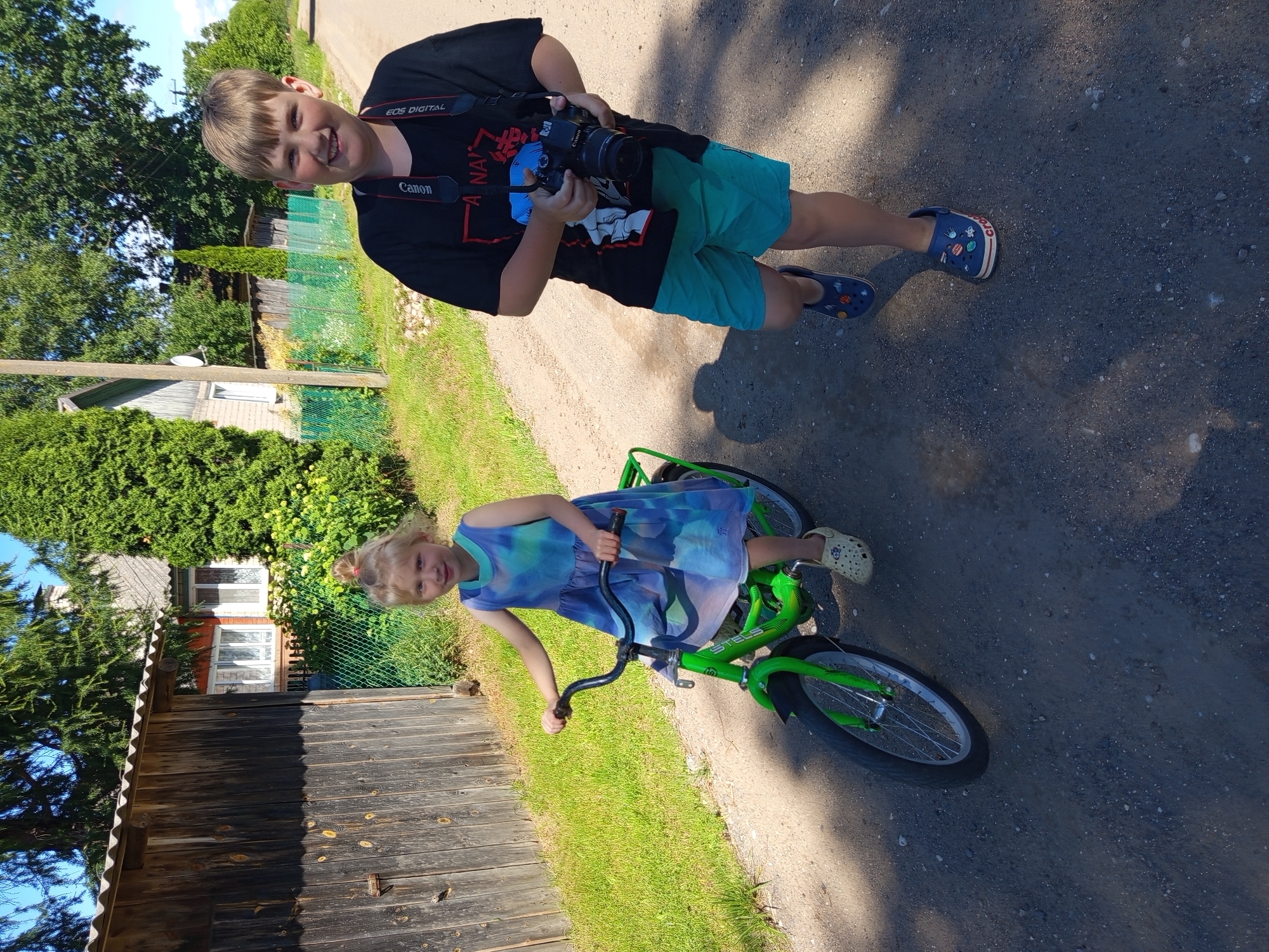 In Ostrovtsy, Russia, a young boy, possibly pre-teen, stands on a dirt path, holding a black Canon digital camera around his neck. He wears a black t-shirt with graphic print and turquoise shorts, along with blue Crocs decorated with colorful charms. He smiles broadly, looking directly at the camera. Beside him, a younger girl, likely a toddler, sits on a bright green bicycle. She wears a blue and green tie-dye dress and cream-colored Crocs. Her hair is tied back in a small bun, and she looks towards the camera with a slight smile. The scene is set outdoors on a sunny day, with lush green grass and trees surrounding the dirt path. A wooden fence and parts of a house are visible in the background. The time of day appears to be midday due to the bright sunlight and distinct shadows. The overall atmosphere is cheerful and relaxed, suggesting a casual outdoor activity. Text visible on the boy's camera strap reads "Canon" and "EOS DIGITAL." The bicycle frame also has the brand name "STELS" visible.