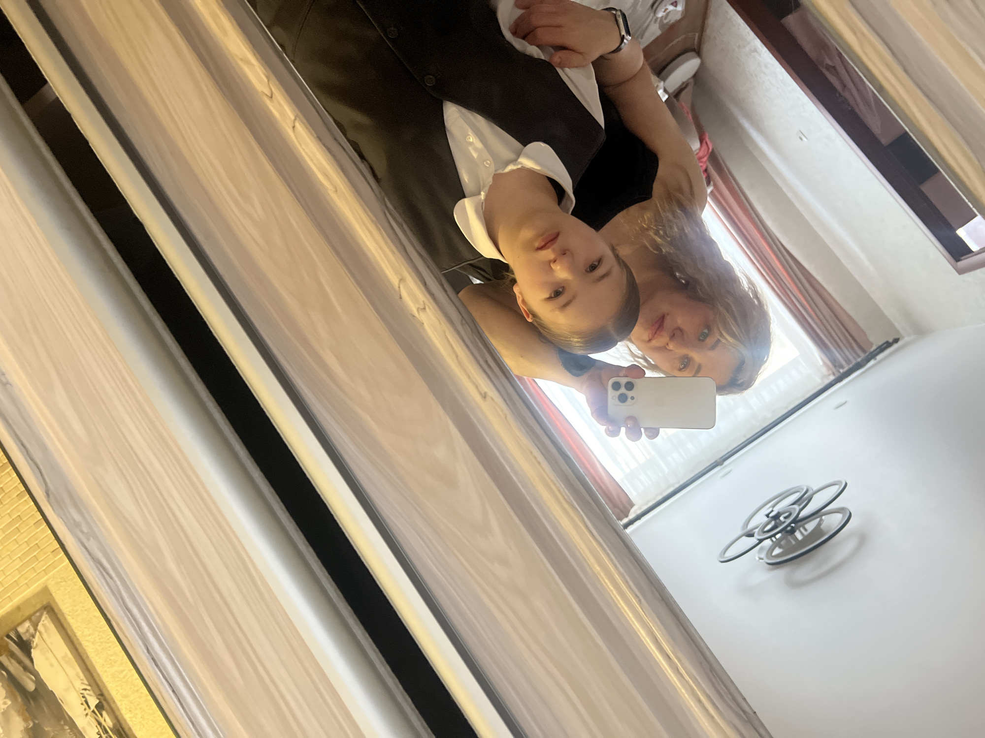 Two females take a mirror selfie inside an interior space with a chandelier on the ceiling. The location is Chaikovsky, Russia. One female is wearing a white collared shirt and a black vest. The second female is wearing a black shirt. Both females are Caucasian. One female is holding a white smartphone, presumably taking the picture. A light-colored door or wardrobe with vertical panels is visible in the reflection. A window with curtains is also reflected.