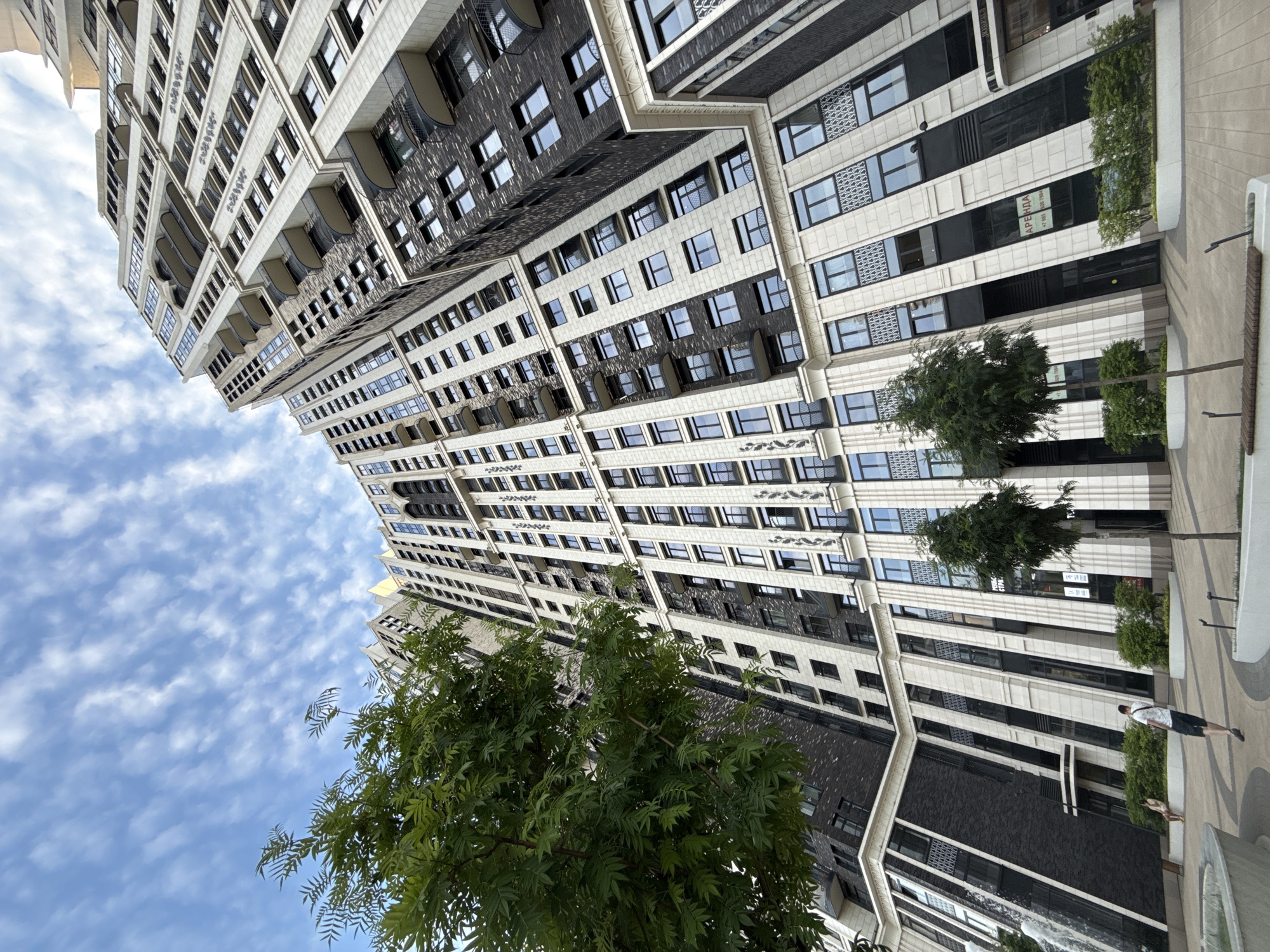 An eye-level, low-angle, outdoor, full-shot captured on a sunny day shows a modern, tall residential building with a complex, angled facade in Moscow, Russia. The building is predominantly composed of white and dark grey materials, featuring numerous windows and balconies. A large green tree is prominently positioned in the foreground on the left, partially obscuring the building's facade. In the mid-ground, two smaller trees are visible. A person in a white t-shirt and dark shorts is walking away from the camera towards the building. Another person is seated in the background to the left, near a fountain feature that includes a cascading water effect. The sky above is blue with scattered white clouds. The ground is paved with light-colored tiles. The overall scene conveys an urban residential environment.
