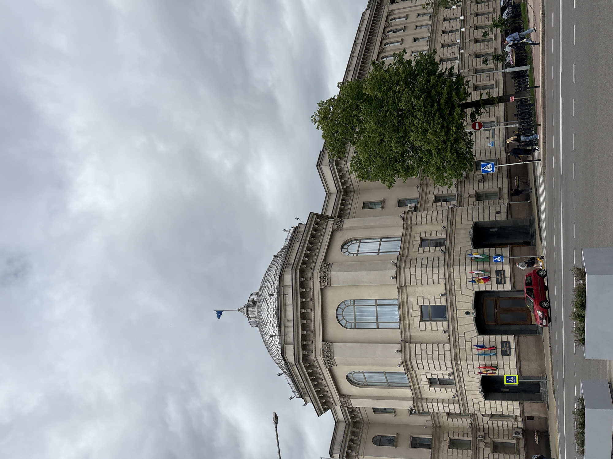 The image shows a building in Minsk, Belarus. The building is made of stone, and it has a dome and a flag on top. There are several flags displayed near the building's entrance along with a red car parked in front of it. A crosswalk is visible with people crossing the street. A street lamp is also present. The sky is overcast and cloudy.