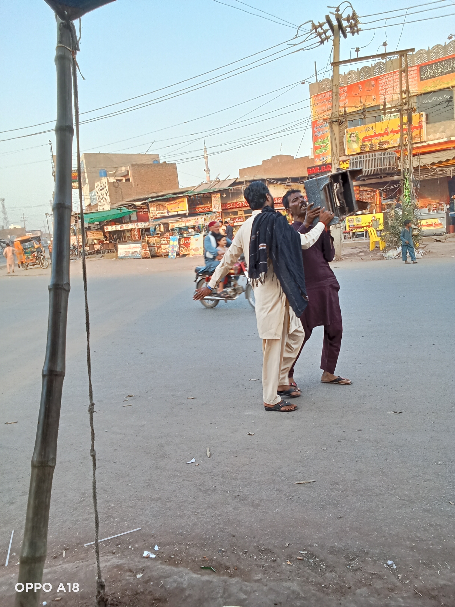 Here is a detailed description of the image:
Two men are standing in the middle of a street, holding a box. The man on the left is wearing a light tan shalwar kameez with a black shawl draped over his shoulder. He is wearing brown sandals. The man on the right is wearing a maroon shalwar kameez and also wearing brown sandals.
In the background, there is a street with shops on either side. There are people walking and riding motorbikes. There is a bamboo pole on the left side of the image. A motorcycle is passing behind the two men. Several people and vehicles are scattered around the street. There is a building with several signs and posters. The sky is clear. The setting appears to be a street scene in a busy commercial area.