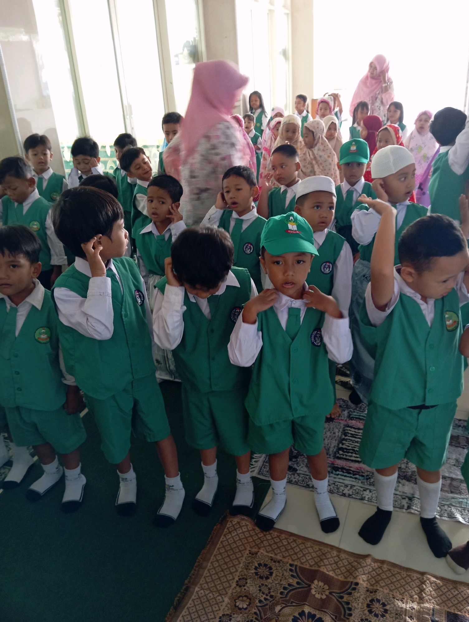 In Malang, Indonesia, a group of young boys, dressed in matching green vests and white shirts, stand together in what appears to be a classroom or communal area. They are likely students at an Islamic school given the attire and the presence of several women in the background wearing hijabs. The boys are mostly looking in the same direction, some with curious expressions, others appearing thoughtful. The environment seems bright, with natural light streaming in from large windows. The floor is covered with a patterned rug, and the walls are a light color. The scene captures a moment of quiet observation or anticipation among the children.