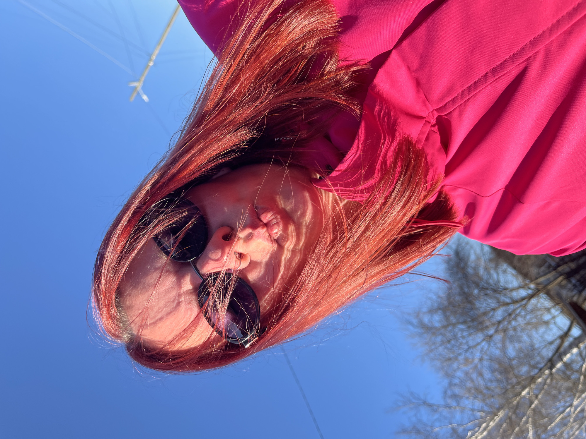 The image is taken from a low angle, looking up at a person with reddish-orange hair and sunglasses. The person is wearing a bright pink jacket. The background is a clear blue sky, with a few bare tree branches visible in the lower right corner. There are also some indistinct structures, possibly utility lines, in the upper left corner. The location is possibly in the city of Chaikovsky, Russia.