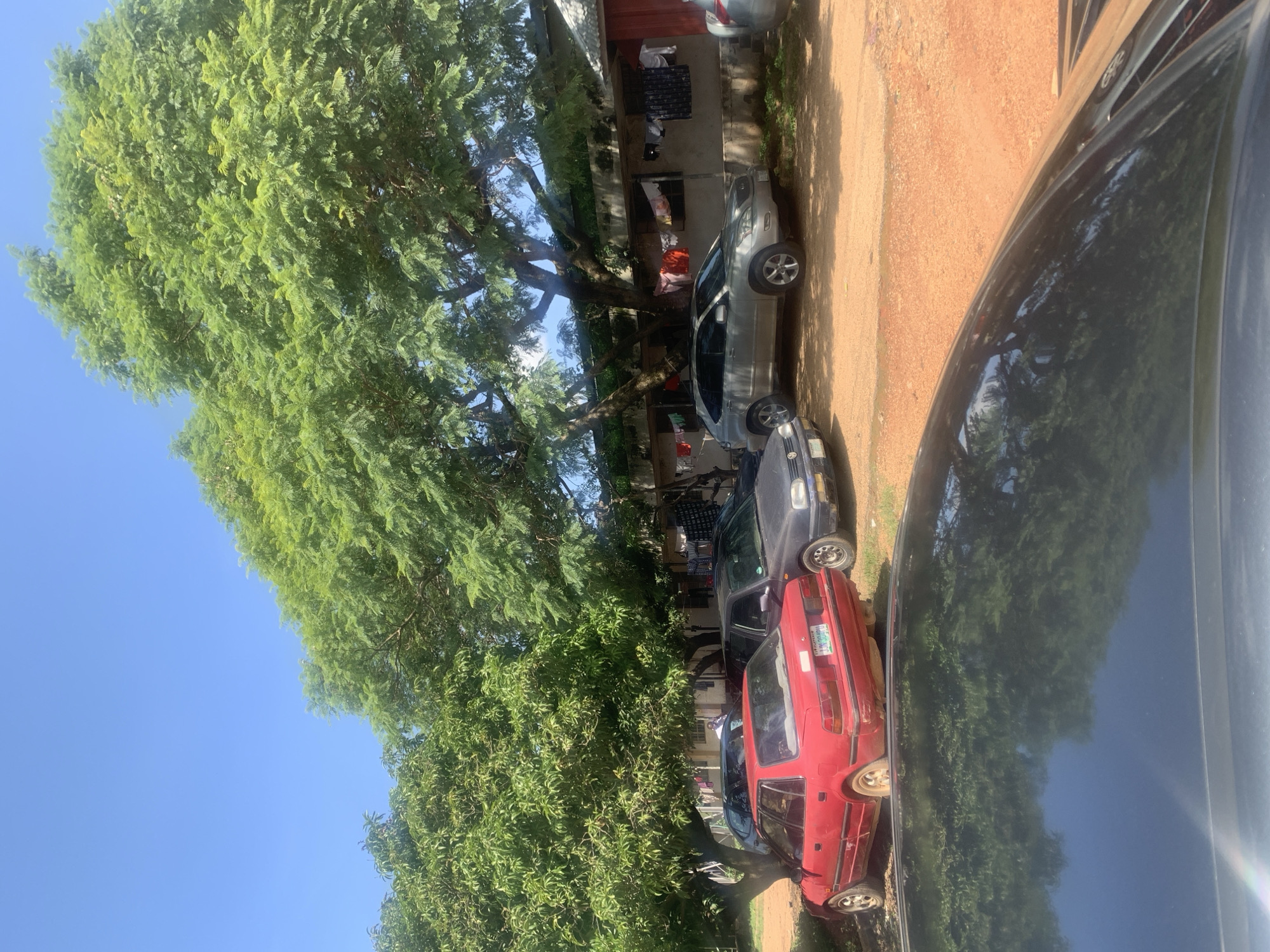 In Jos, Nigeria, on a sunny day, a red sedan with a license plate reading "JIN 304 ADH" is parked on a dirt lot next to a black sedan. Behind them, a silver SUV is parked in front of a building where laundry hangs on a clothesline. The scene is shaded by the large, leafy branches of a tree. In the foreground, the reflection of trees can be seen on the dark, glossy surface of a car's hood.
