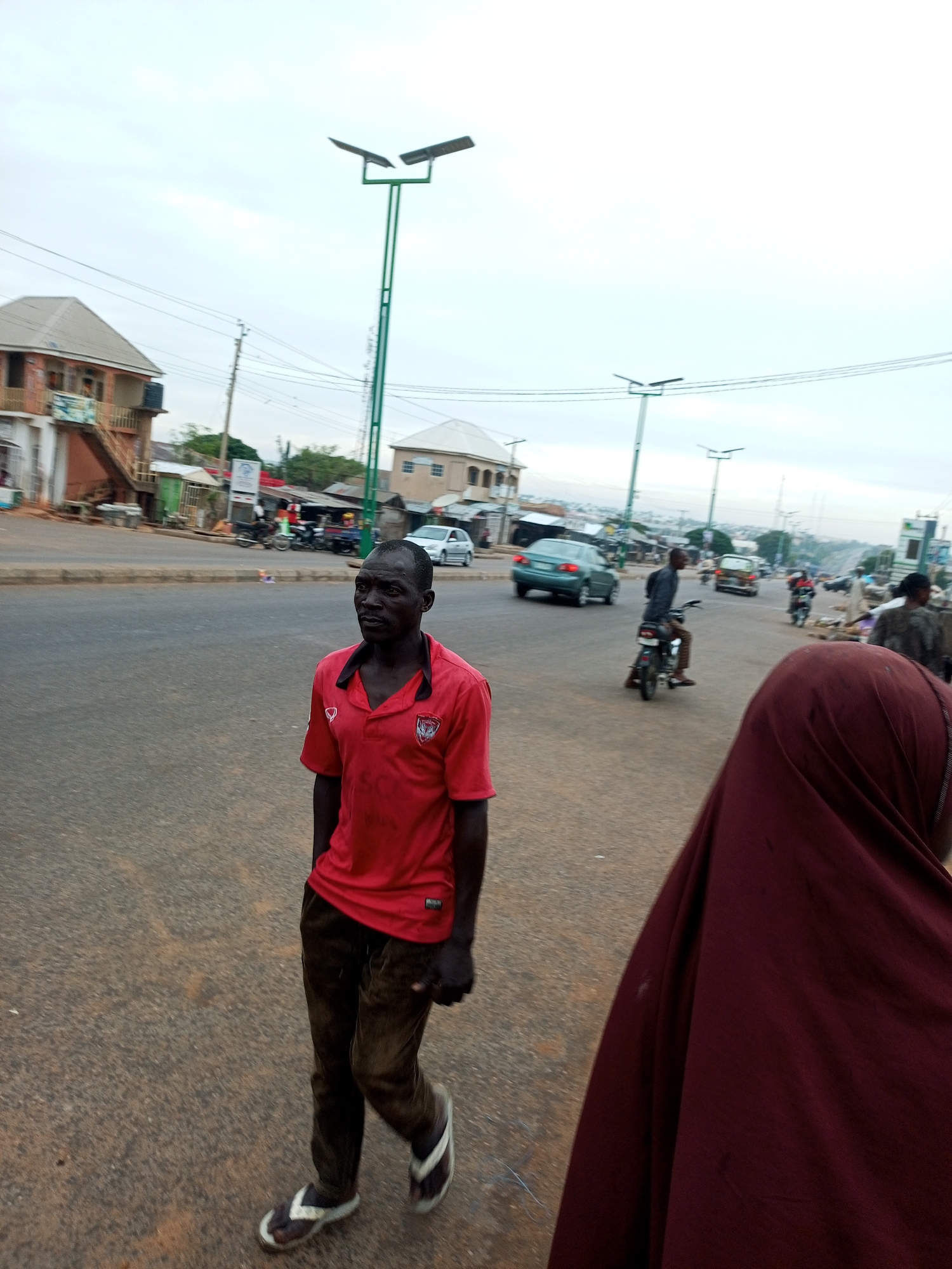 A medium shot captures an overcast day on a street in Tempure, Nigeria. In the foreground, a man walks towards the camera, dressed in a red polo shirt and dark pants. He wears flip-flops. To the right and slightly behind him, a person wearing a burgundy hijab is visible. The street is paved and appears to be a main thoroughfare with traffic. In the background, buildings, streetlights, and other vehicles, including a motorcycle and cars, are present. The overall scene depicts a typical urban environment with everyday activities.