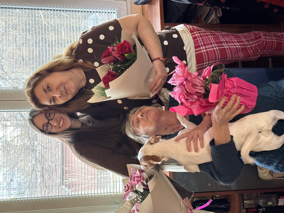 A forensic description of the image reveals an indoor celebratory scene in a residence located in Sofia, Bulgaria. Three females and a small dog are depicted.
To the left, a younger female with long dark hair and glasses, wearing a brown sweater, smiles directly at the camera while holding a bouquet of pink orchids wrapped in light paper.
To the right, a middle-aged female with brown hair, wearing a brown polka-dot short-sleeved top and red plaid pajama pants, also smiles at the camera, holding a bouquet of red roses with baby's breath, wrapped in light paper.
In the center foreground, a seated older female with grey hair, wearing a dark blue sweater, holds a small white and brown dog in her left arm. Her eyes are closed, and her head is tilted back slightly. Her right hand supports a pink potted cyclamen plant, wrapped in pink polka-dot paper. The dog faces away from the camera, looking towards the older woman's face.
The background features a large window with horizontal white blinds, through which diffused daylight illuminates the room. Outside, faint tree branches are visible. To the right of the subjects, a dark wooden desk or cabinet is partially visible. To the left, a shelving unit holds several wine bottles and other items. The overall lighting suggests a daytime setting.