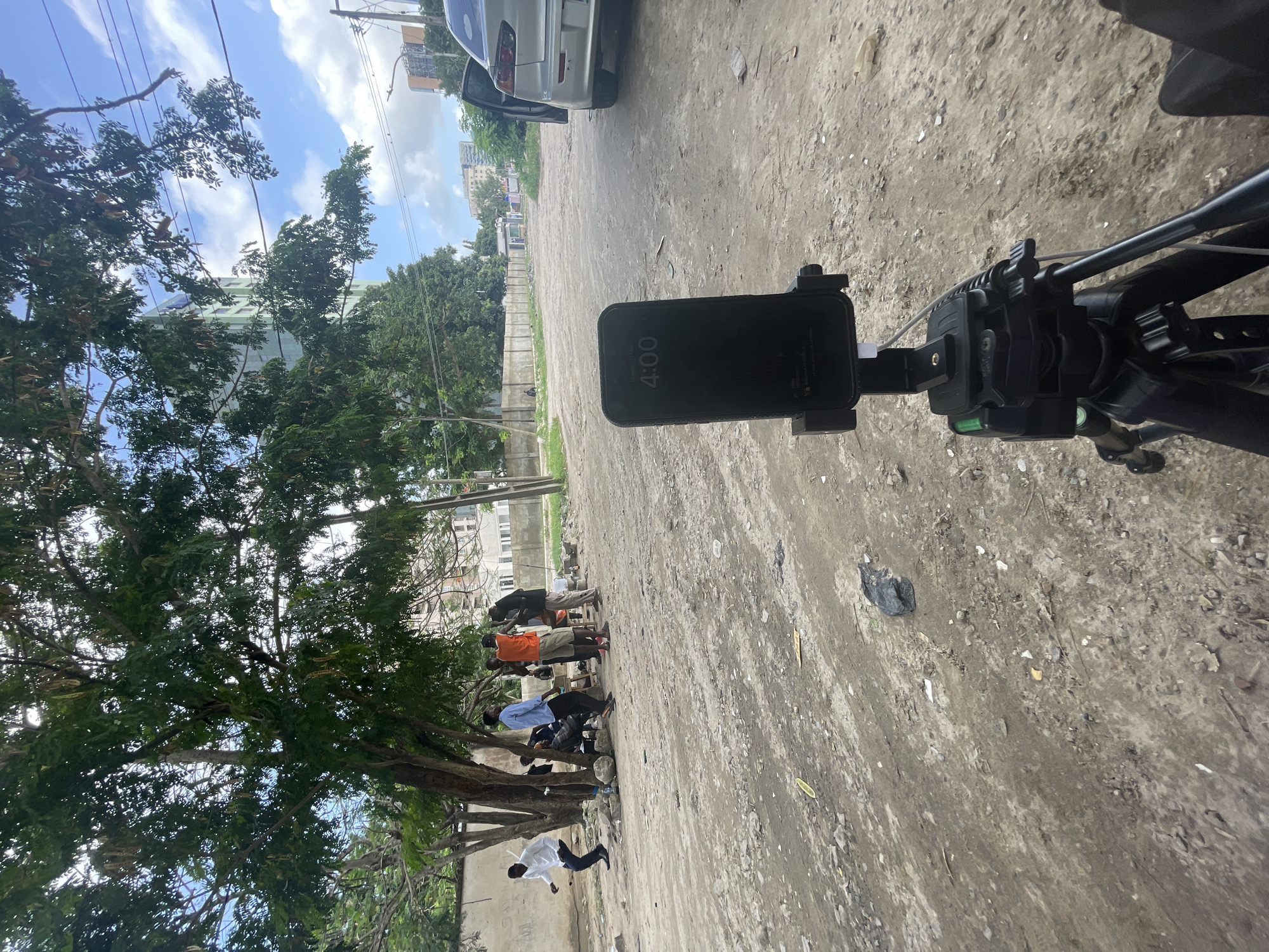 The image shows an iPhone mounted on a tripod in an outdoor location in Ikoyi, Nigeria. The phone's screen displays the time as 4:00. The surrounding environment is a partially paved area adjacent to a street with buildings in the background. A group of people is visible near a tree, seemingly engaged in various activities. A silver car is parked nearby. The ground appears uneven and consists of dirt, gravel, and scattered debris. The overall environment suggests an urban or suburban setting.