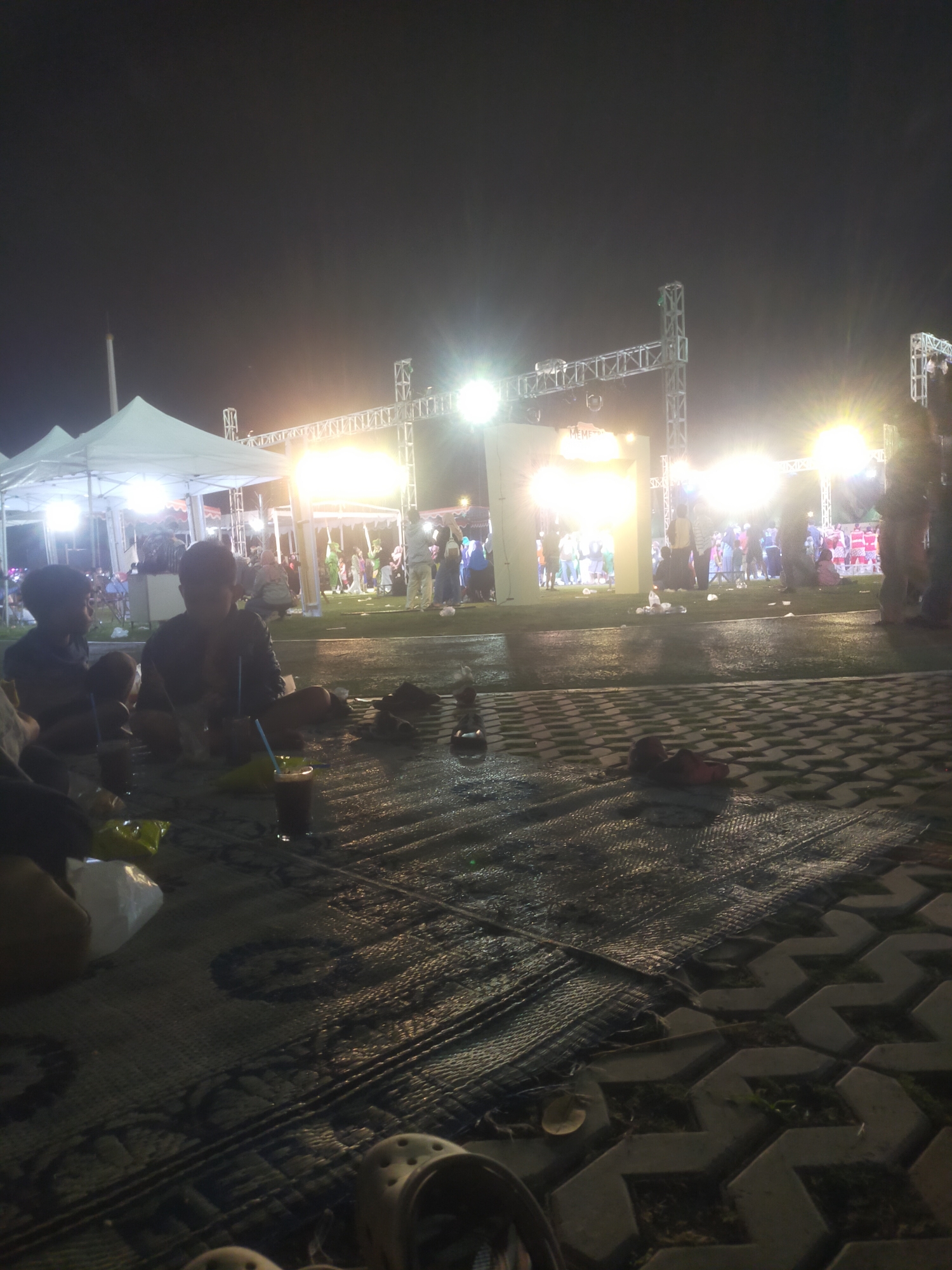 The image captures an outdoor evening scene, likely at a public event or festival, in Bantul, Indonesia. In the foreground, a group of individuals are seated on mats on the ground. Two young boys are visible, one with dark hair and the other appearing to be a toddler. There are drinks in glasses and some food packaging on the mats. The ground appears wet, possibly from recent rain.
In the midground and background, a brightly lit stage structure is set up, adorned with scaffolding and numerous lights, illuminating the scene. A crowd of people is gathered in front of the stage, some standing and others seated. Several white tents are also visible, suggesting vendors or information booths. The overall atmosphere appears festive and lively, with the bright lights contrasting with the dark night sky. The paving stones in the lower right corner of the image are a distinctive feature of the paved area.