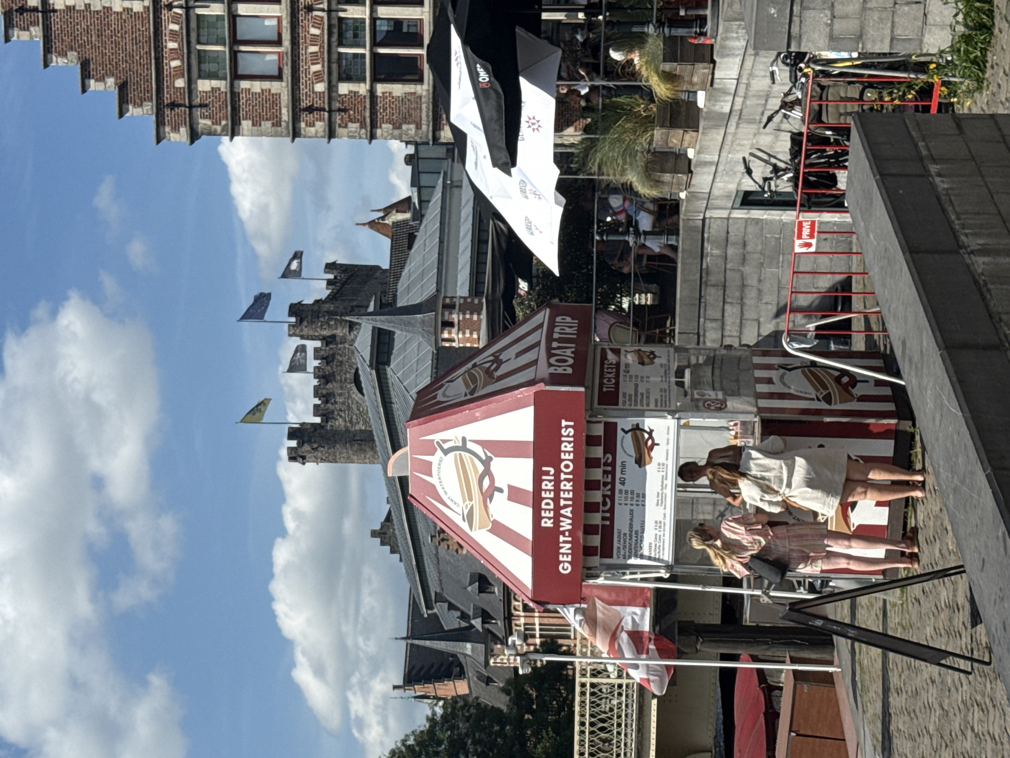 The image captures a sunny day in Ghent, Belgium, with a view of the Graslei and Korenlei canal area. In the foreground, a red and white striped booth for "REDERIJ GENT-WATERTOERIST" (Ghent Water Tours) is prominently displayed, offering boat trips. Two women are seen interacting at the ticket counter, one in a striped dress and the other in a cream-colored dress. A third woman, likely an employee, stands behind the counter. Adjacent to the booth, a chalkboard sign stands open. In the background, the medieval Gravensteen castle, with its imposing stone structure and flags, is visible against a sky filled with white clouds. Historic brick buildings with stepped gables characteristic of Ghent's architecture line the canal. Several people are seated at tables under umbrellas on a terrace to the right of the boat tour booth. Bicycles are parked behind a red metal barrier further to the right, with a sign that reads "PRIVE" (private). The cobblestone pavement and the stone embankment suggest a well-trodden tourist area.