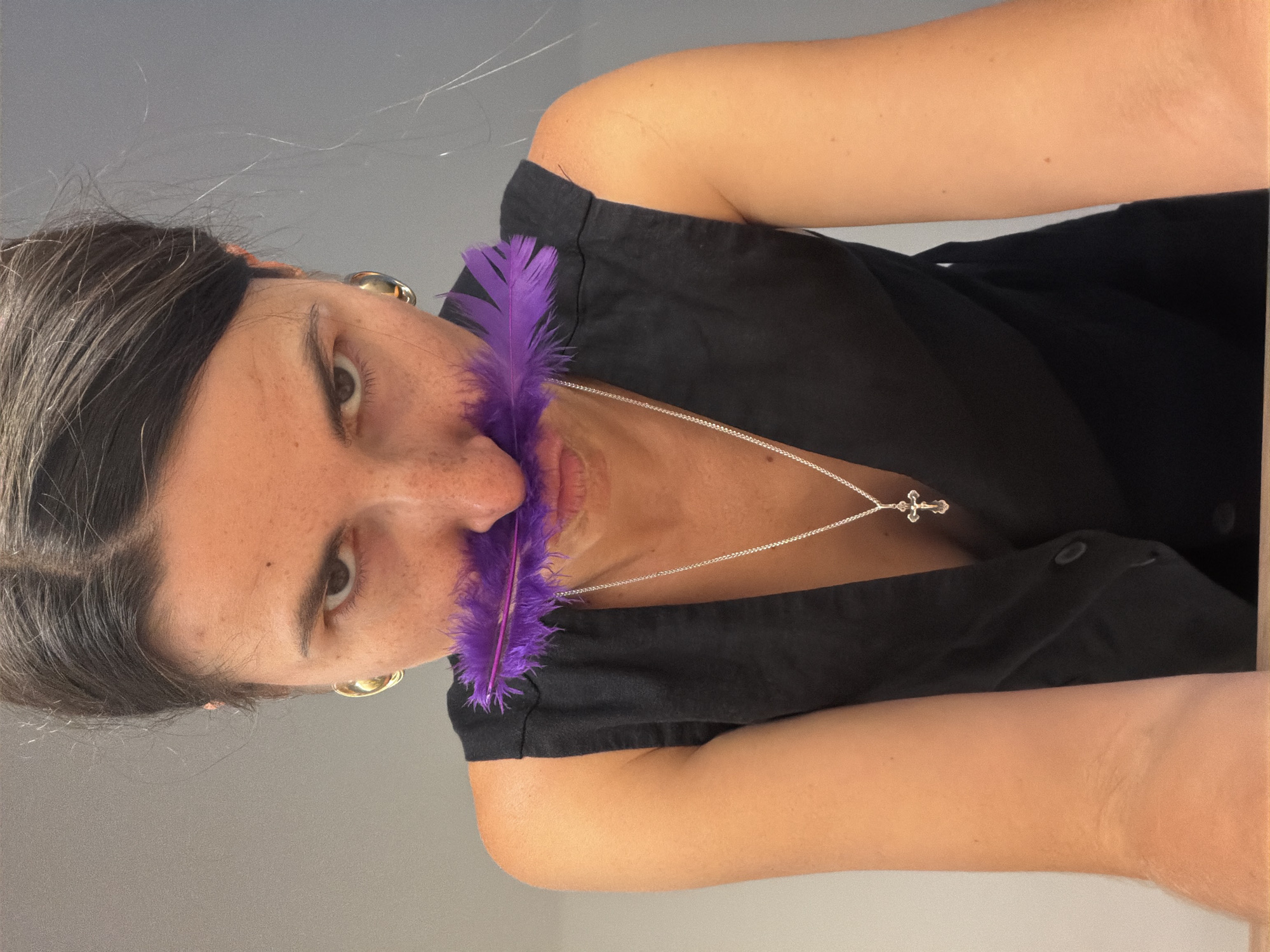 A close-up, portrait-style shot features a person with dark hair and freckles, looking directly at the viewer with a neutral expression. The image is rotated 90 degrees counter-clockwise, showing the subject from an overhead perspective, with their head to the left and body extending to the right. A vibrant purple feather is held close to their nose and mouth.
The person is wearing a black sleeveless top and a silver chain necklace with a small silver cross pendant. A gold hoop earring is visible on their right ear. Their dark hair, with some visible lighter strands, is pulled back. The skin tone is light to medium, with noticeable freckles across the face.
The activity captured is a posed shot, with the feather as a central prop, drawing attention to the subject's face. The background is a plain, light gray wall, suggesting an indoor setting. There are no specific time of day or weather cues, and no visible text in the image. The scene is located in Budva, Montenegro.