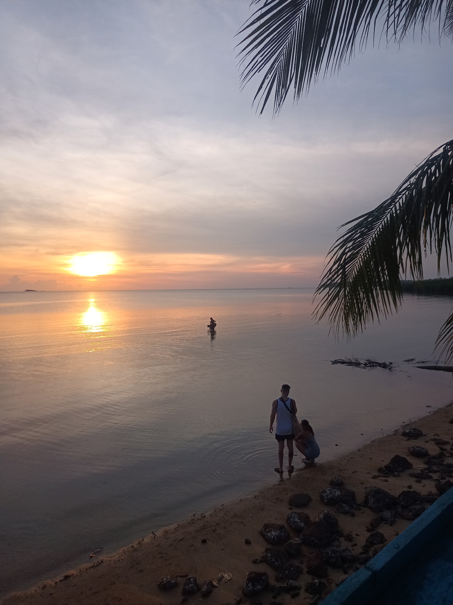 The image captures a serene scene at Baan Wok Tum, Thailand, during sunset. The sun is setting over a calm body of water, with its reflection shimmering on the surface. Two individuals are standing on the sandy beach at the water's edge; a man is standing while the other person is crouching near the shoreline. A palm tree's fronds frame the upper part of the image, adding a tropical touch. The beach is a mix of sand and rocks. Another person is seen wading out in the water further out.