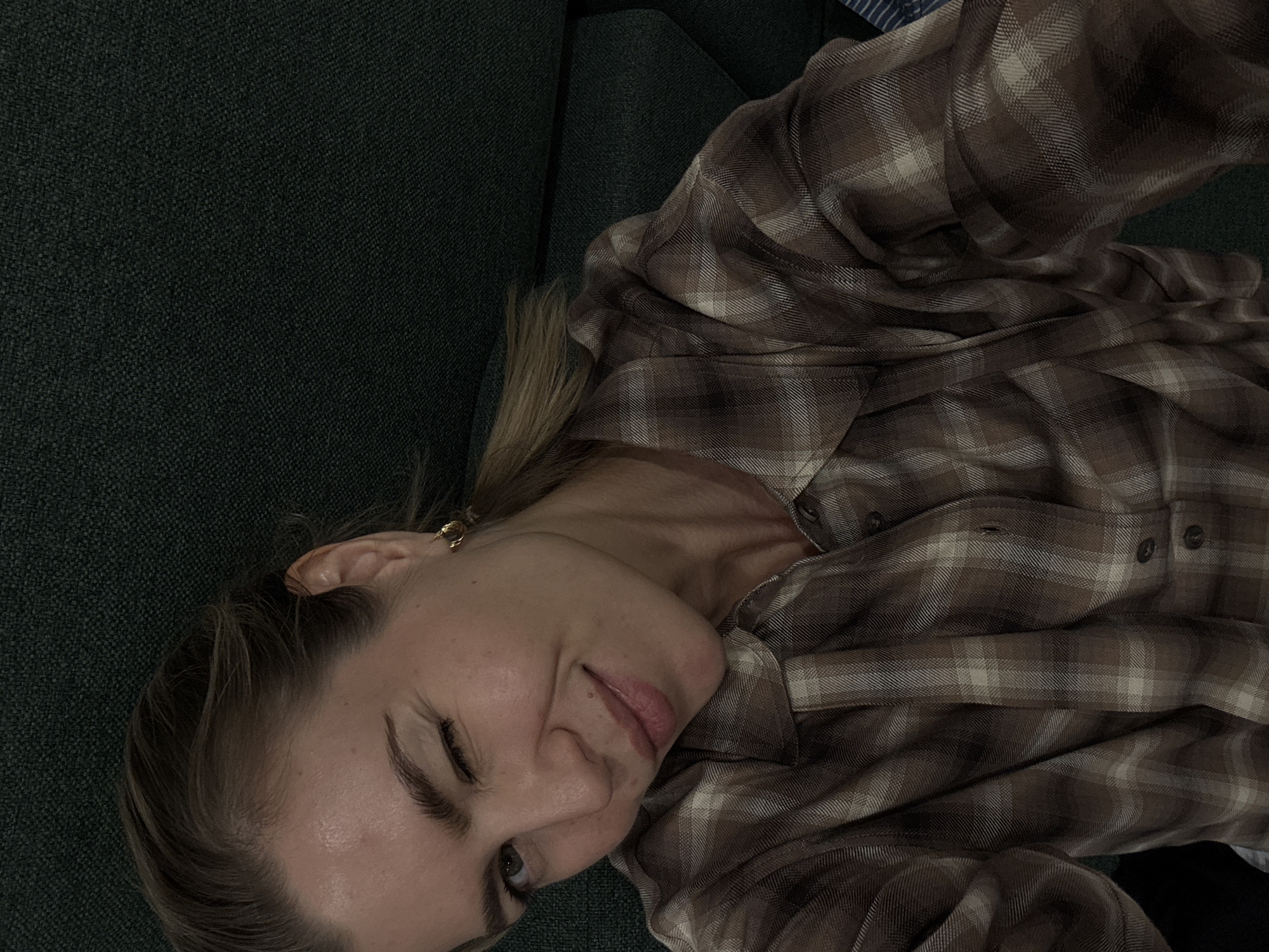 A woman with blonde hair and light skin is lying on a dark green couch or other soft surface. She is wearing a brown and white plaid shirt. She is facing the camera with a slight smile. Her head is tilted to the left. A small gold hoop earring is visible in her left ear. The location is unknown.