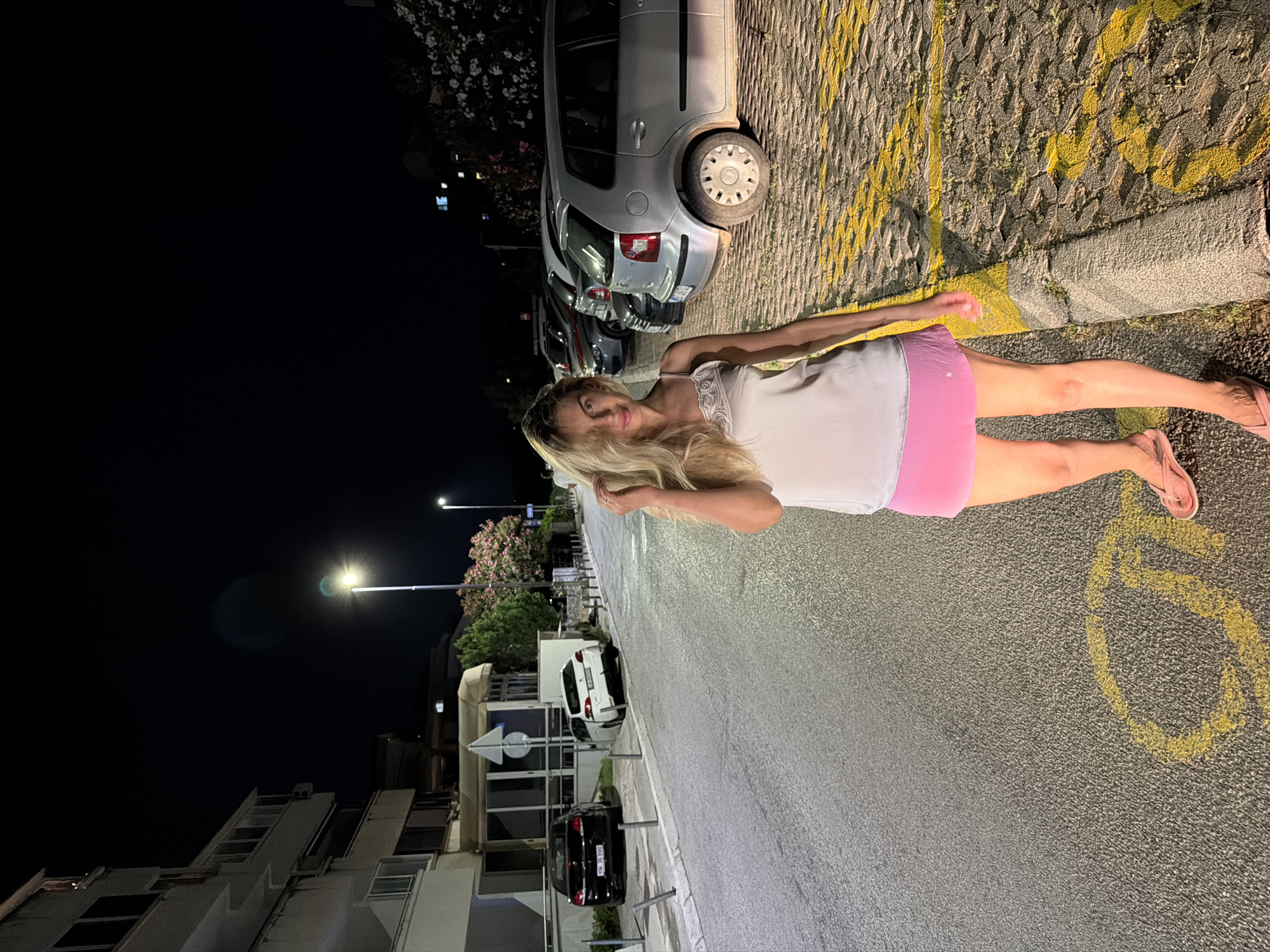 A woman stands on a street at night in Budva, Montenegro. She is wearing a light-colored camisole and a pink skirt, with pink flip-flops on her feet. Her hair is long and blonde. The street is paved and has yellow parking markings. In the background, there are parked cars and a building. A street light illuminates the scene.