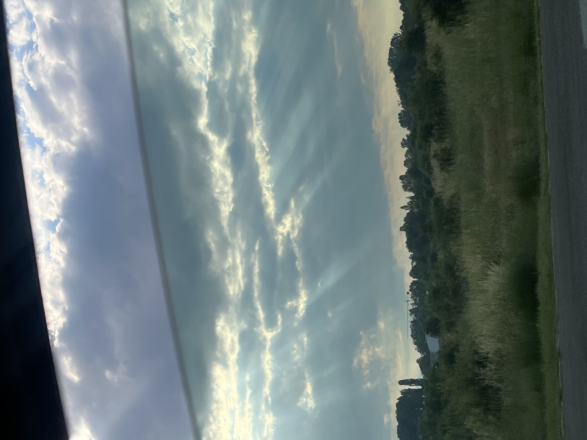 The image shows a landscape view through a window, with the sky dominating the upper portion. Dramatic light rays, often referred to as crepuscular rays, are visible piercing through a cloudy sky, suggesting a sunrise or sunset. Below the sky, a dense line of trees and vegetation stretches across the horizon. In the distance, a white building and a construction crane are visible among the trees. The foreground features a patch of tall grass and a section of asphalt road, indicating an outdoor setting, possibly a roadside view. The overall atmosphere is serene and natural, with the play of light and shadow creating a visually striking scene. The location is stated as Unknown, Unknown.