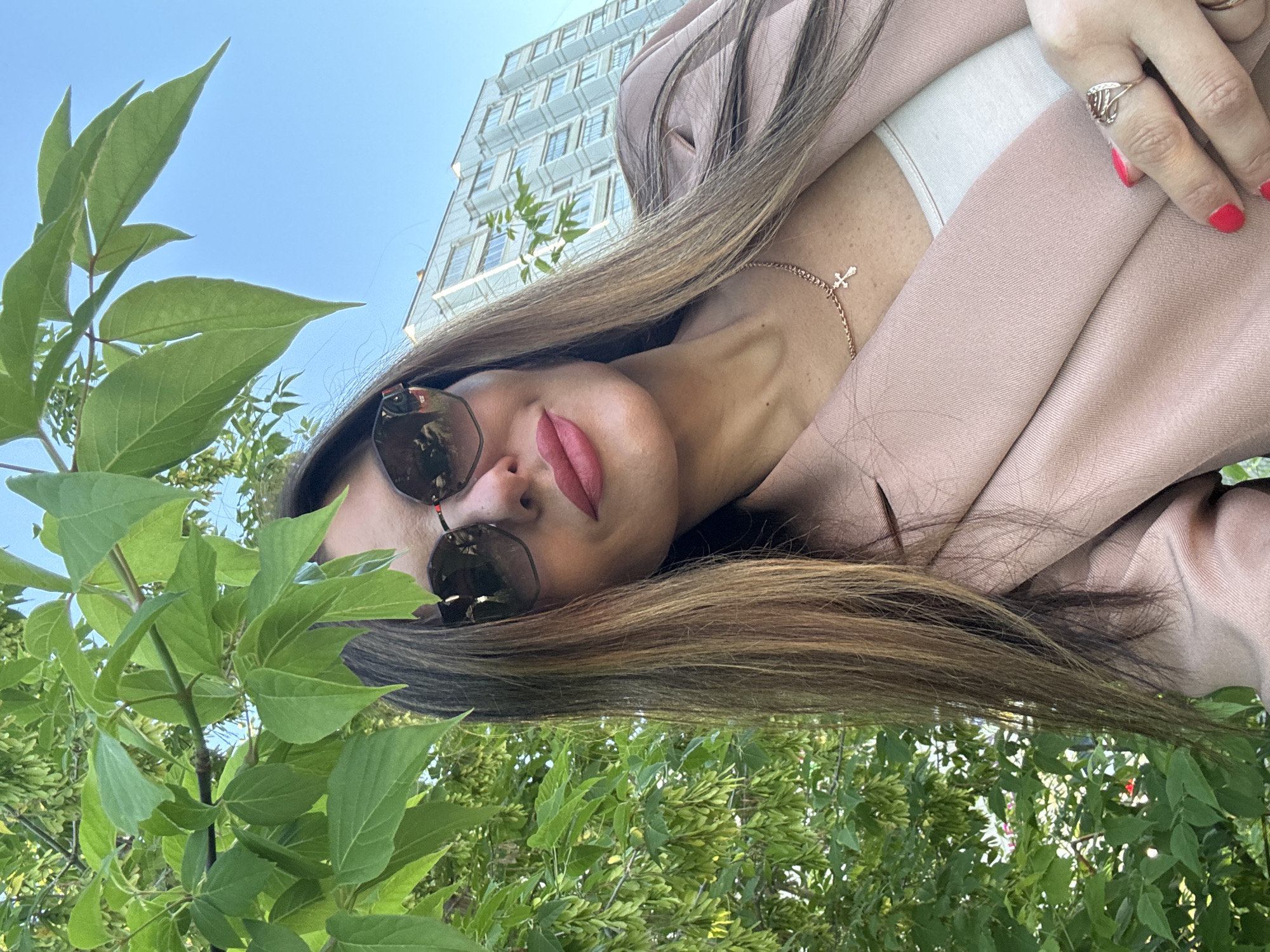 A close-up, eye-level selfie shows a fair-skinned woman with long, wavy brown hair, wearing oversized sunglasses, a light pink blazer, and a white top. She has on a rose gold chain necklace with a small cross pendant and a rose gold ring with a filigree design on her right ring finger. Her lips are painted a bright pink. The woman is positioned in front of lush green foliage, with a light blue sky and a modern apartment building in the background. The photo was taken on a sunny day, likely in the city of Chaikovsky, Russia.