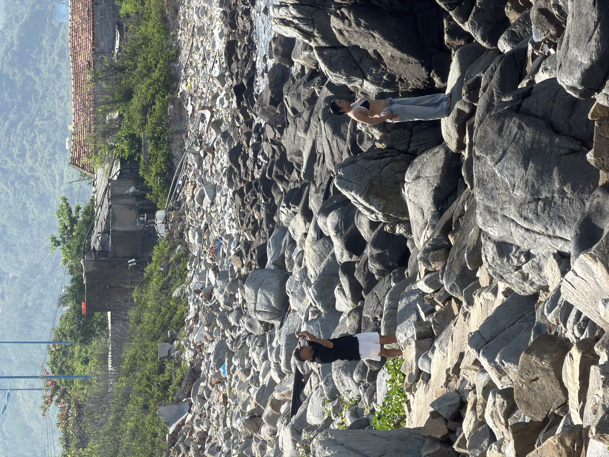 The image depicts a rocky shoreline in Vĩnh Lương, Vietnam. In the foreground, a man wearing a black t-shirt and white shorts stands on a rock, holding a phone to take a photo. To the right, a woman in a bikini top and light-colored pants stands on another rock, facing away from the camera. The shoreline is covered in various sizes of grey and brown rocks, with some green foliage growing among them. In the background, there are some small buildings with tiled roofs and a lush green hillside. The sea is visible in the distance, with calm water reflecting the sunlight.