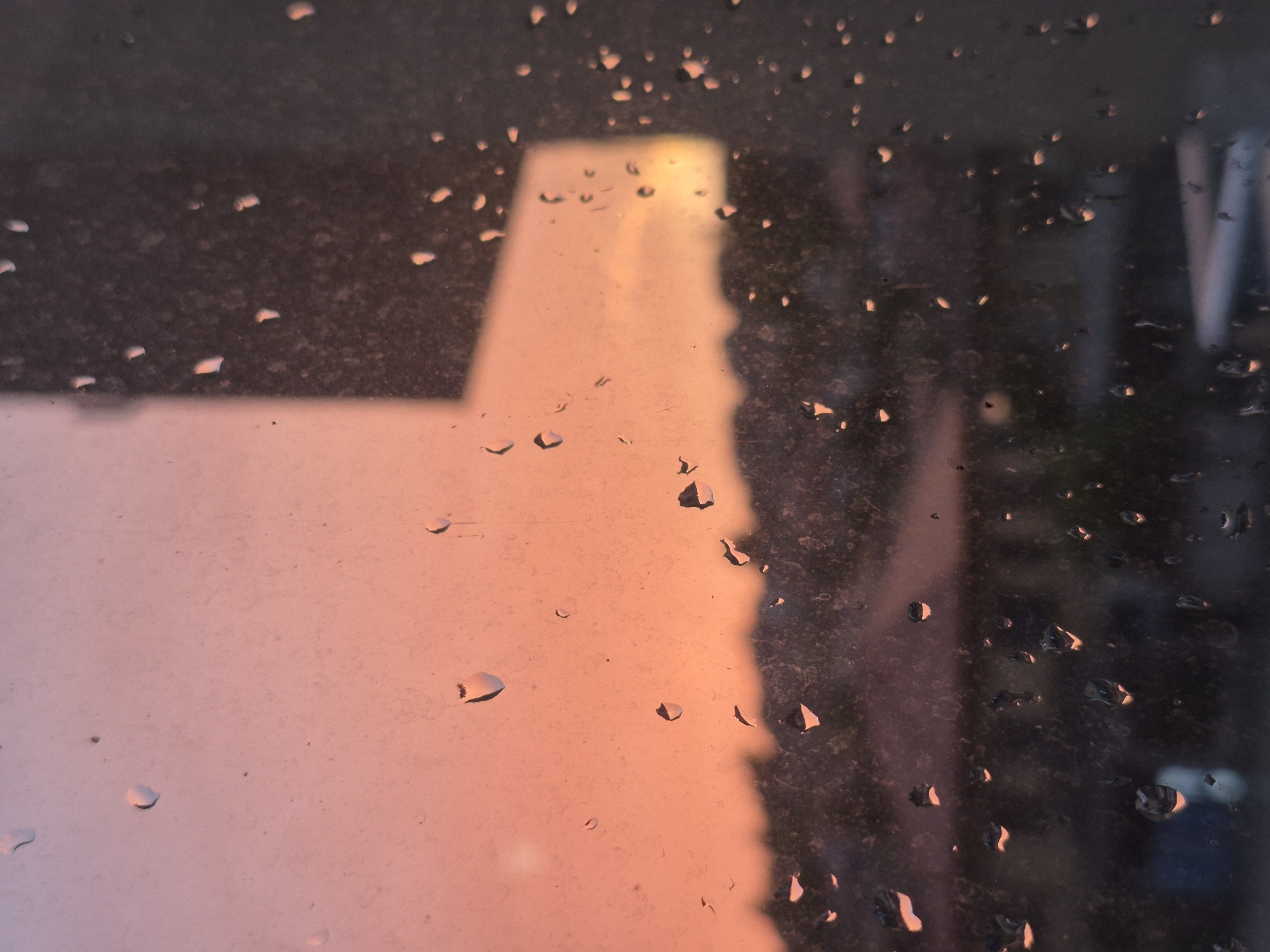 The image is a close-up, low-angle shot of a window with raindrops on it, viewed from inside. The raindrops distort the view of the exterior scene, which appears to be a city at sunset or sunrise. The sky is a gradient of orange and pink, with a blurred silhouette of buildings or trees in the foreground. The overall mood is calm and reflective. The specific location in Popovo, Bulgaria, is not discernible from the image.