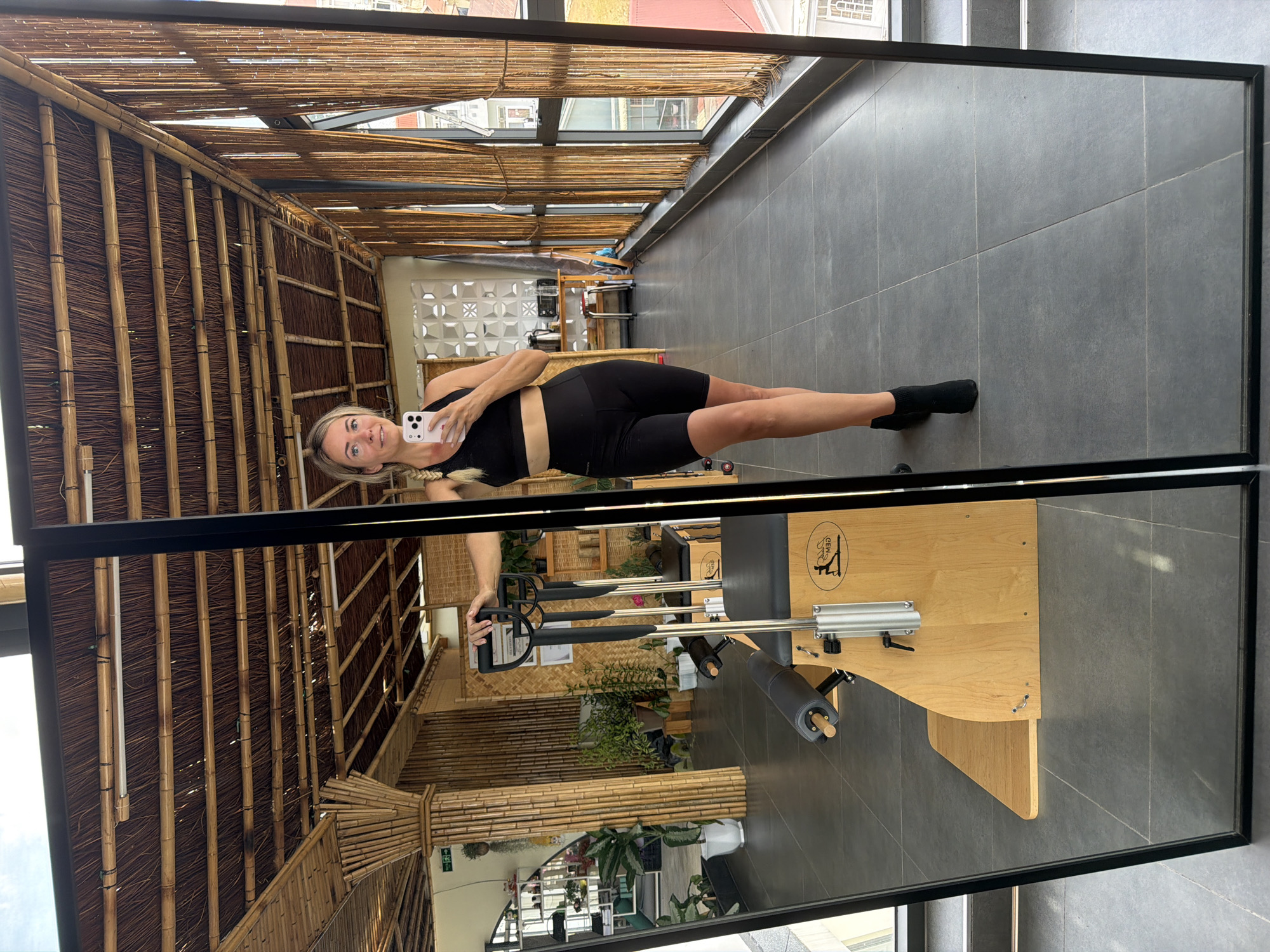 A blonde woman in athletic wear takes a mirror selfie in a Pilates studio in Da Lat, Vietnam. She is wearing a black sports bra and black bike shorts, with black ankle boots. The studio features a bamboo ceiling and walls, with large windows offering a view of the city. In the reflection, a Pilates reformer machine is visible, along with shelves stocked with plants and exercise equipment. The floor is tiled in a dark gray. The woman is holding her phone with her right hand, and her left hand is resting on the reformer.