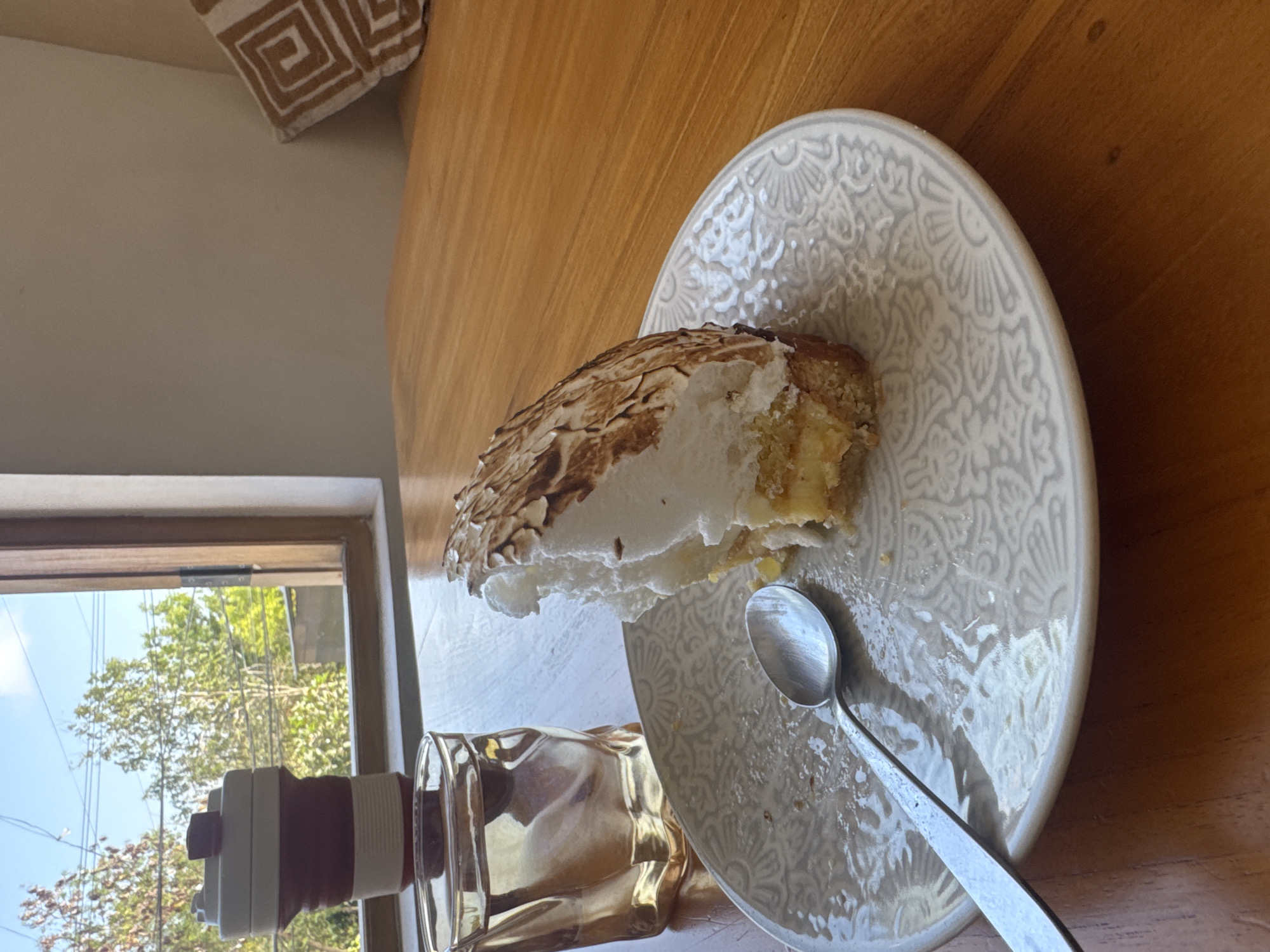 A close-up, low-angle shot shows a slice of lemon meringue pie on a grey, ornate plate. The pie has a golden-brown toasted meringue topping, a yellow filling, and a crumbly crust. A silver spoon rests on the plate, slightly to the left of the pie. To the left of the plate is a clear glass with a yellowish liquid, possibly water or a light-colored drink. Behind the glass and plate is a white and red collapsible coffee cup. The setting appears to be near a window, with blurred trees and power lines visible through the glass. The table is made of light-colored wood. A brown and white patterned pillow is visible in the background on the right. The scene is likely indoors, in a cafe or restaurant setting in Kuta Selatan, Indonesia.