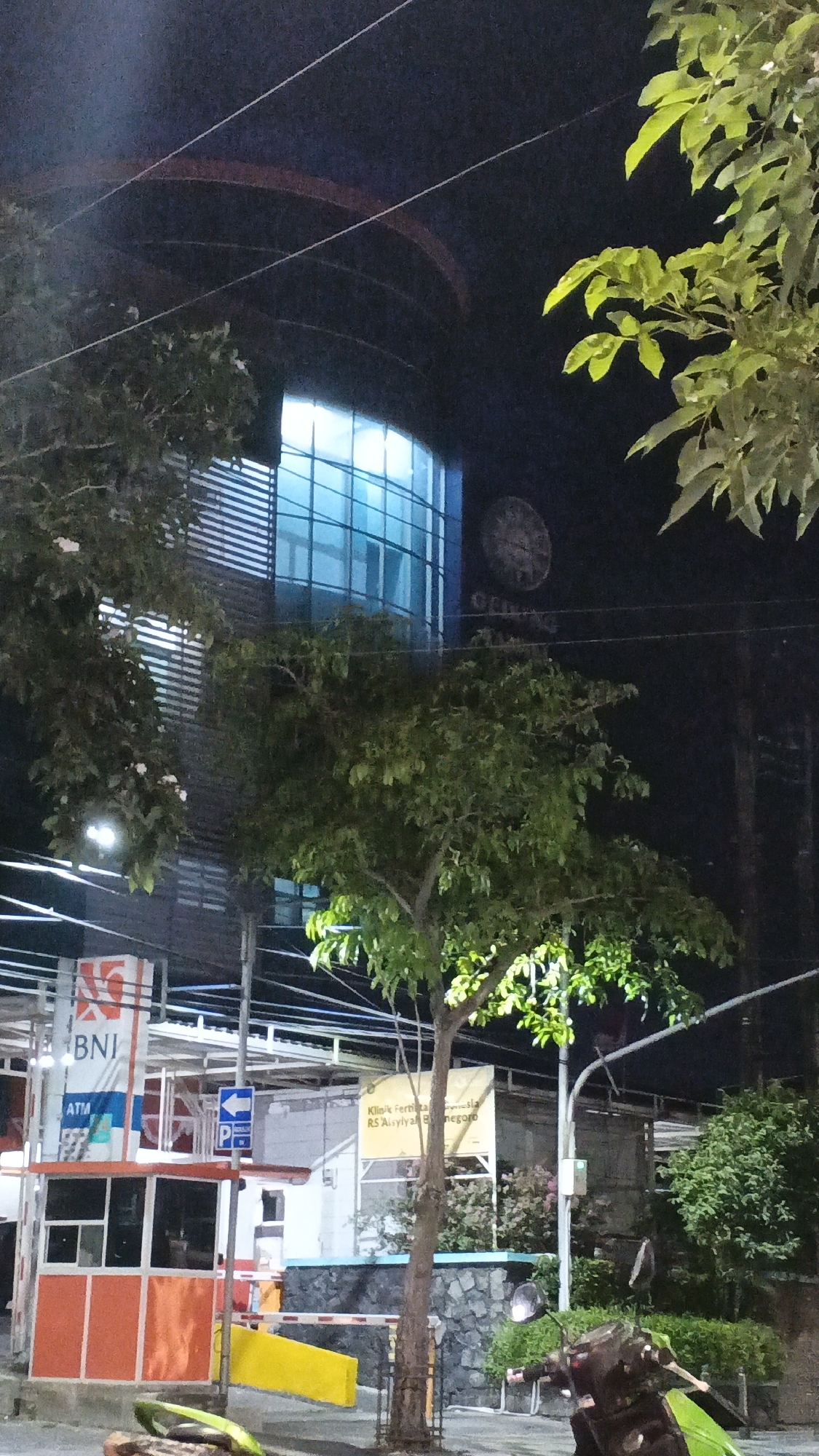 The image depicts an outdoor nighttime scene in Bojonegoro, Indonesia, likely near a hospital or clinic. A multi-story building with a curved facade featuring blue glass windows is visible in the background. A large tree with lush green foliage stands in the foreground, partially obscuring the building. To the left, a guard booth with orange and white panels and a sign for "BNI" and "ATM" indicates a financial institution or service point. A blue directional arrow sign with a "P" symbol suggests parking is available. Further to the right, another sign reads "Klinik F**nita RS Aisyiyah B**negoro," confirming the presence of a clinic and hospital. A motorcycle is parked on the right side of the image, and a yellow barrier is visible across the road. The overall environment appears to be urban, with streetlights and overhead wires present.