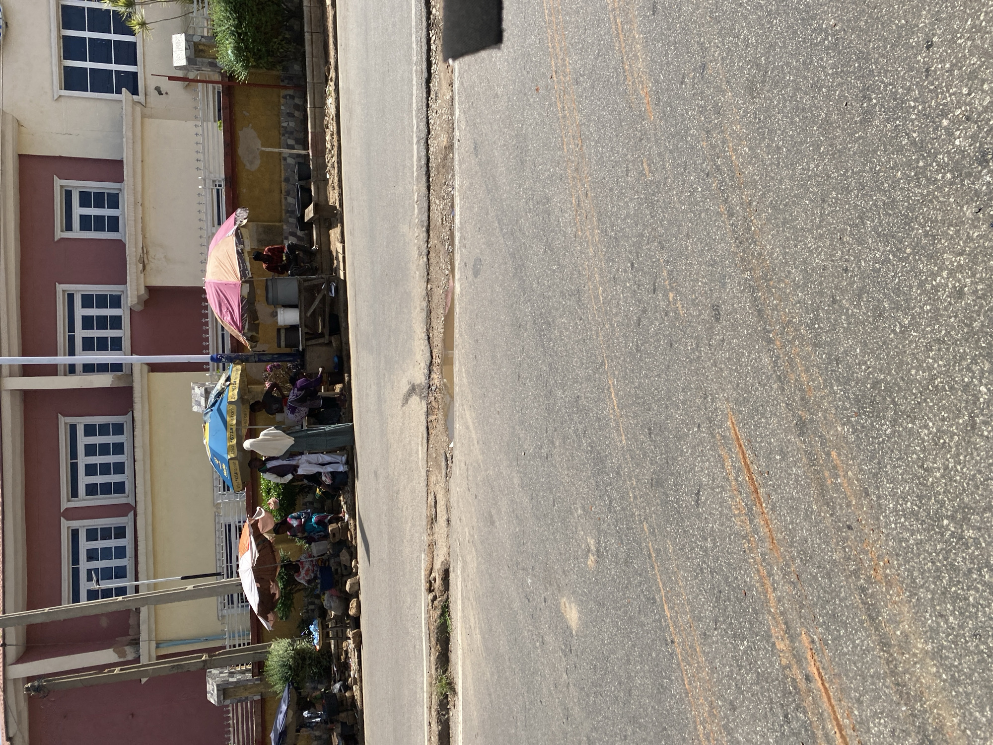 A street scene in Jos, Nigeria, features a building with a reddish-pink facade and cream-colored sections, adorned with large, white-framed windows. In front of the building, several individuals are gathered around market stalls. Two umbrellas, one with orange and white stripes and another with pink and yellow stripes, provide shade. People are seen standing and sitting, some appearing to be vendors and others customers. Various objects, likely merchandise or supplies, are visible on tables and the ground. A paved road runs in front of the market area, with some patches of grass and debris along the edge. The overall impression is a busy, informal marketplace situated alongside a main thoroughfare.