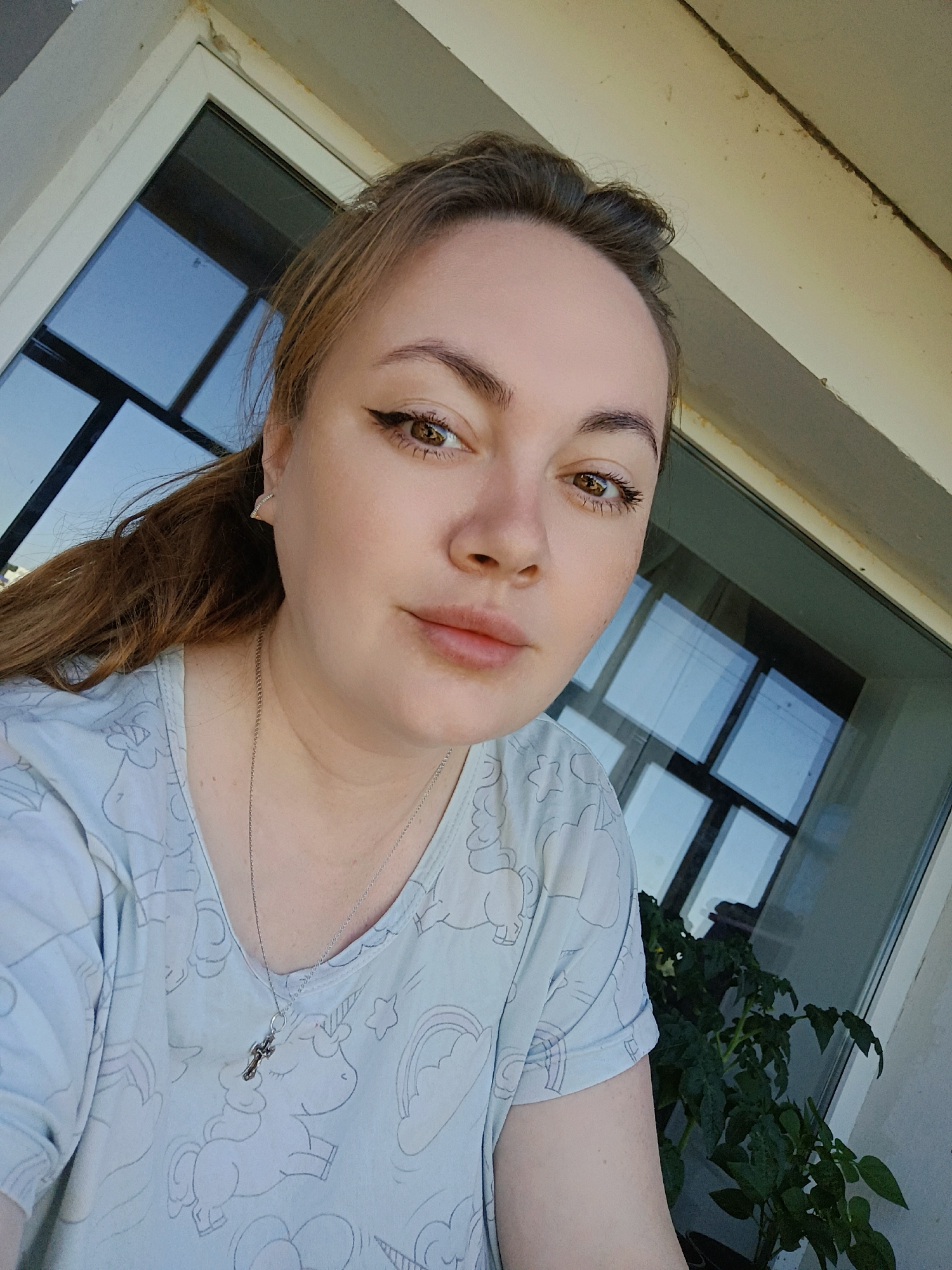 A young Caucasian woman with light brown hair pulled into a ponytail is pictured in a close-up selfie taken outdoors. She is wearing a light blue t-shirt with a pattern of unicorns and clouds. A silver chain with a cross pendant is visible around her neck. She has winged eyeliner and appears to be looking at the camera. The background shows a window with reflections of the sky and a potted plant with dark green leaves. The architecture suggests a building located in the city of Chaikovsky, Russia.