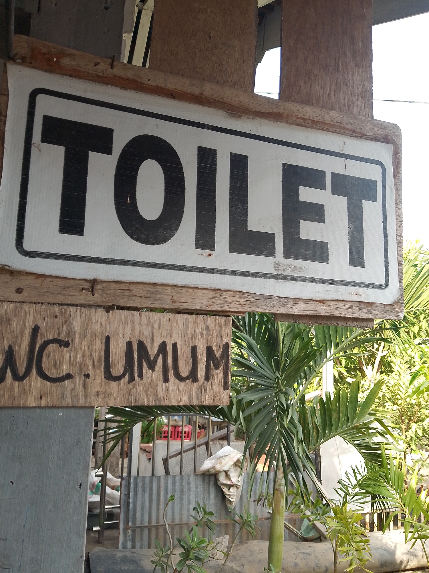 The image shows two signs. The upper sign, framed by wood, reads "TOILET" in large black capital letters against a white background. Below it, another wooden plank sign has handwritten text in black that appears to read "WC. UMUM". Behind the signs, a tropical environment is visible, with palm fronds and other greenery. In the background, there are structures made of corrugated metal and some objects that could be trash or supplies. The overall scene suggests an outdoor, possibly rustic, location. Given the text "WC. UMUM" which translates to "Public Toilet" in Indonesian, the location is likely a public restroom facility in Indonesia. Based on the provided context that the image is from Teep, Indonesia, this scene is depicting an outdoor public restroom in Teep, Indonesia.