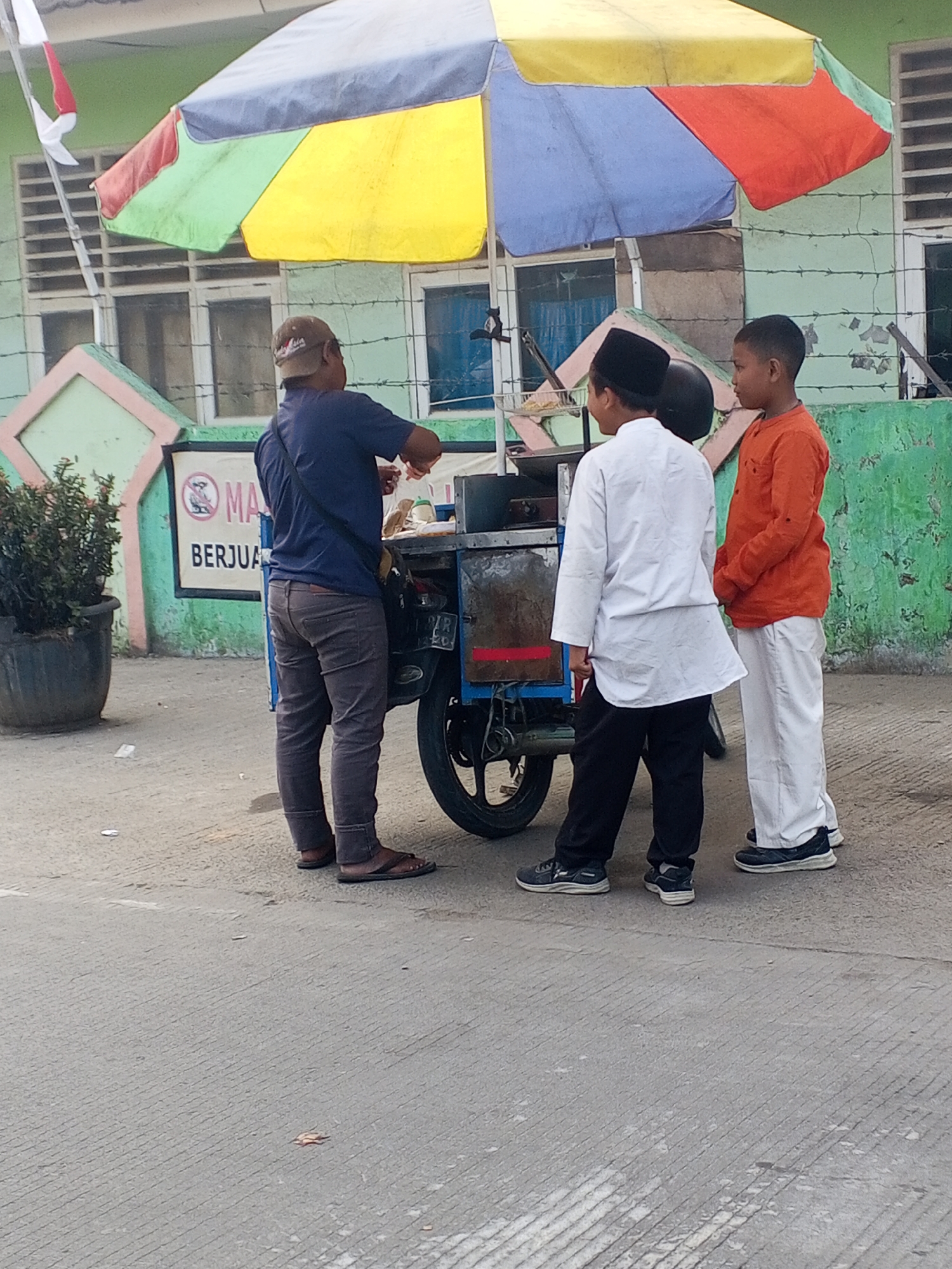 In front of a weathered green building with a pink and green trimmed facade, three individuals stand near a food cart under a colorful umbrella. The building has barred windows. A man wearing a dark blue t-shirt, brown pants, and flip-flops stands behind the food cart, appearing to serve or prepare food. Two young boys, one in a white tunic and black pants, and the other in an orange t-shirt and white pants, stand to the side of the cart. The ground is paved with concrete. A potted plant is visible to the left of the scene, and a red and white Indonesian flag is fluttering on a pole behind the umbrella. The scene appears to be in the city of Ciruas, Indonesia.
