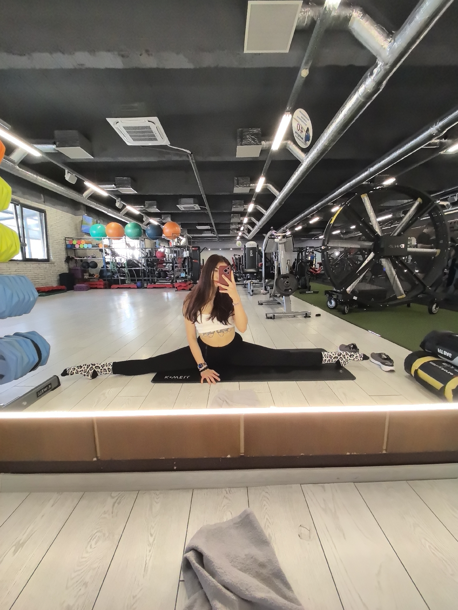 A full-length shot captures a woman in a gym, doing a split on a black mat labeled "K-METT," while holding a cell phone up to take a selfie. She wears a white top and black leggings, along with patterned socks. The reflection in the mirror shows a gray towel on the floor. The gym features fitness balls, weights, and exercise equipment. The location is Stara Zagora, Bulgaria.