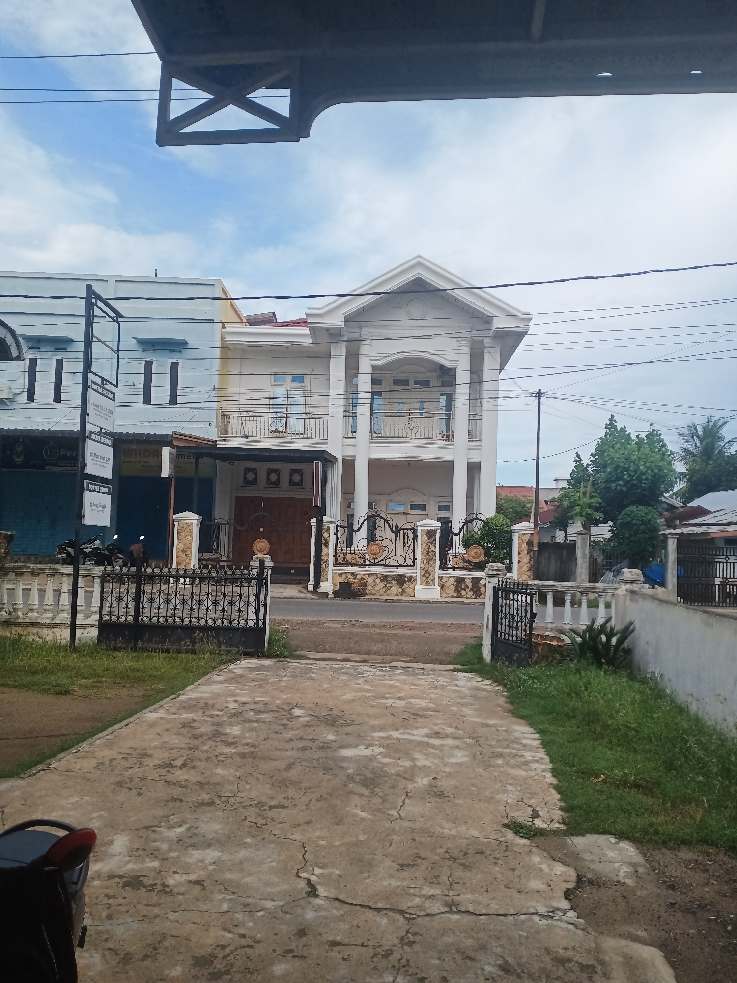 The image shows a street in Lhokseumawe, Indonesia, with several buildings. The main focus is a large, white, two-story house with a prominent triangular pediment on the facade. It has columns and a balcony with an ornate railing. To the left of this house is a light blue, three-story building, likely a commercial or residential property, with multiple windows and a dark awning. Between these two main buildings, there is a smaller section with a dark wooden door and decorative ironwork. A black wrought-iron fence with white balusters lines the front of the properties, separating them from the street. A paved driveway leads towards the houses. Grass and some small plants grow alongside the fence and driveway. Power lines crisscross the sky above. In the foreground, a portion of a black and red motorcycle is visible on the paved surface. The sky is partly cloudy.