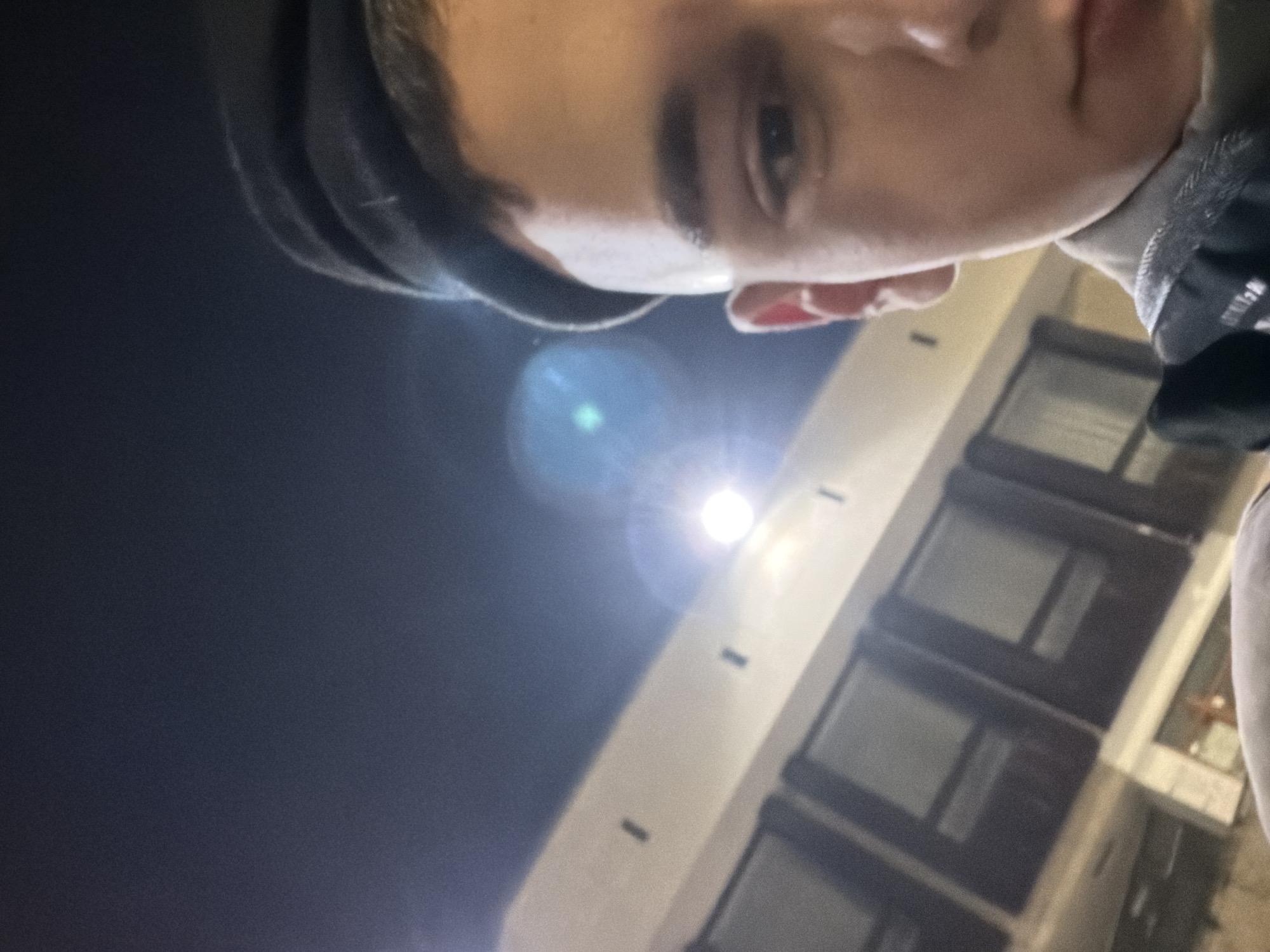 The image appears to be a selfie taken at night in an urban environment. A person with dark hair, wearing a black beanie, is facing the camera from a low angle. The background shows a building with a light-colored facade and dark-framed windows. A bright streetlight creates lens flare in the center. The location is unspecified, and there's no indication of this location being Panagyurishte, Bulgaria, or any particular city. No actions or interactions are evident.
