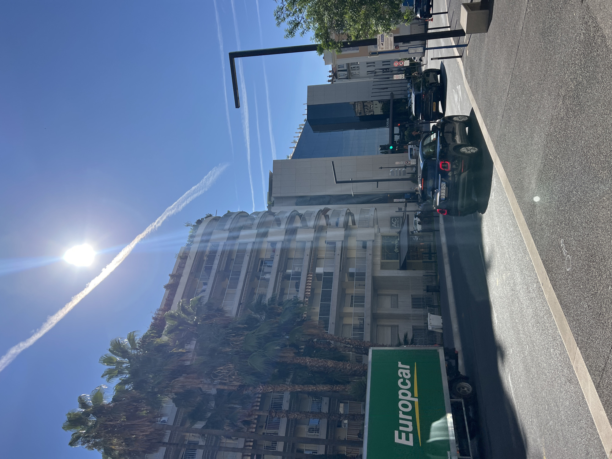 In Nice, France, on a sunny day, a Europcar truck is parked on the left side of the street. In the background, a curved apartment building with balconies and palm trees in front of it stands tall. To the right of the apartment building, a modern glass-fronted building with the "AC HOTELS" logo is visible. Several cars are driving on the street, including a black Fiat 500 in the foreground. The sky is blue with thin white contrails from an airplane. The street is paved with asphalt and bordered by a sidewalk.