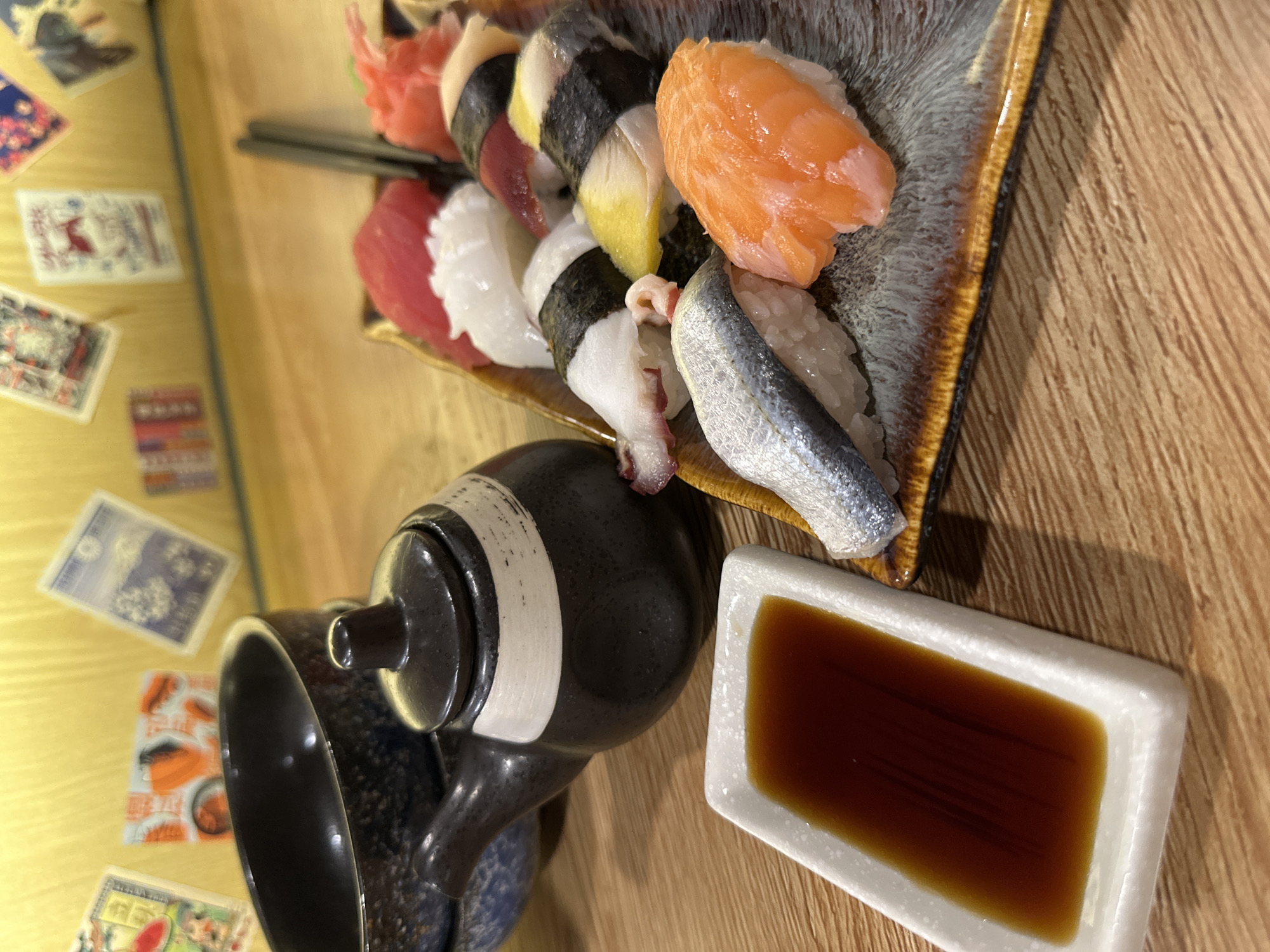 This image captures a close-up view of a plate of assorted sushi, arranged on a textured brown and grey ceramic platter. The sushi appears to include various types of fish, such as salmon and tuna, as well as what looks like octopus and a silver-skinned fish, all atop rice. A small white rectangular dish filled with dark soy sauce is positioned to the right of the sushi. In the lower left, a dark, possibly ceramic teapot with a white band and a textured pattern is partially visible, along with the rim of a larger serving vessel. The sushi and accompanying items are placed on a light brown wooden surface, likely a table. Scattered around the wooden surface are several small, colorful cards with what appear to be Japanese motifs or illustrations. The lighting suggests an indoor setting, possibly a restaurant. The overall impression is that of a prepared meal, ready to be eaten.