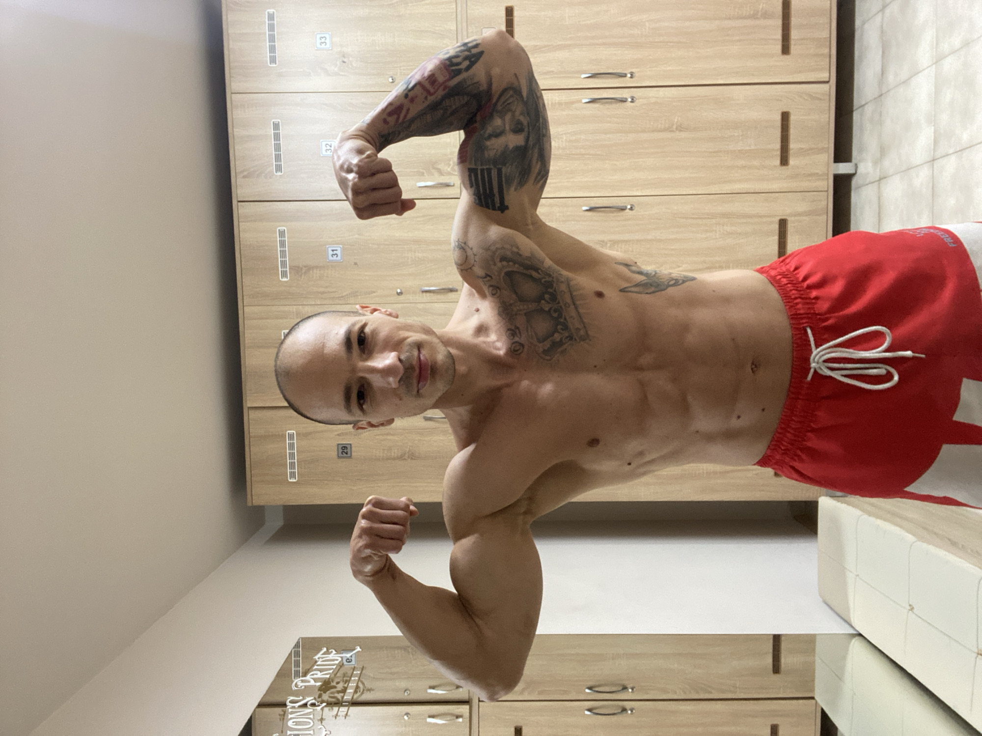 A muscular man with tattoos is posing and flexing his biceps in a locker room. He is wearing red and white shorts. The locker room features light-colored wooden lockers with numbers, a white bench, and neutral-toned walls. The location is inferred to be in Pleven, Bulgaria, based on the presence of "LION'S PRIDE PLEVEN" visible on a reflective surface in the lower left corner.