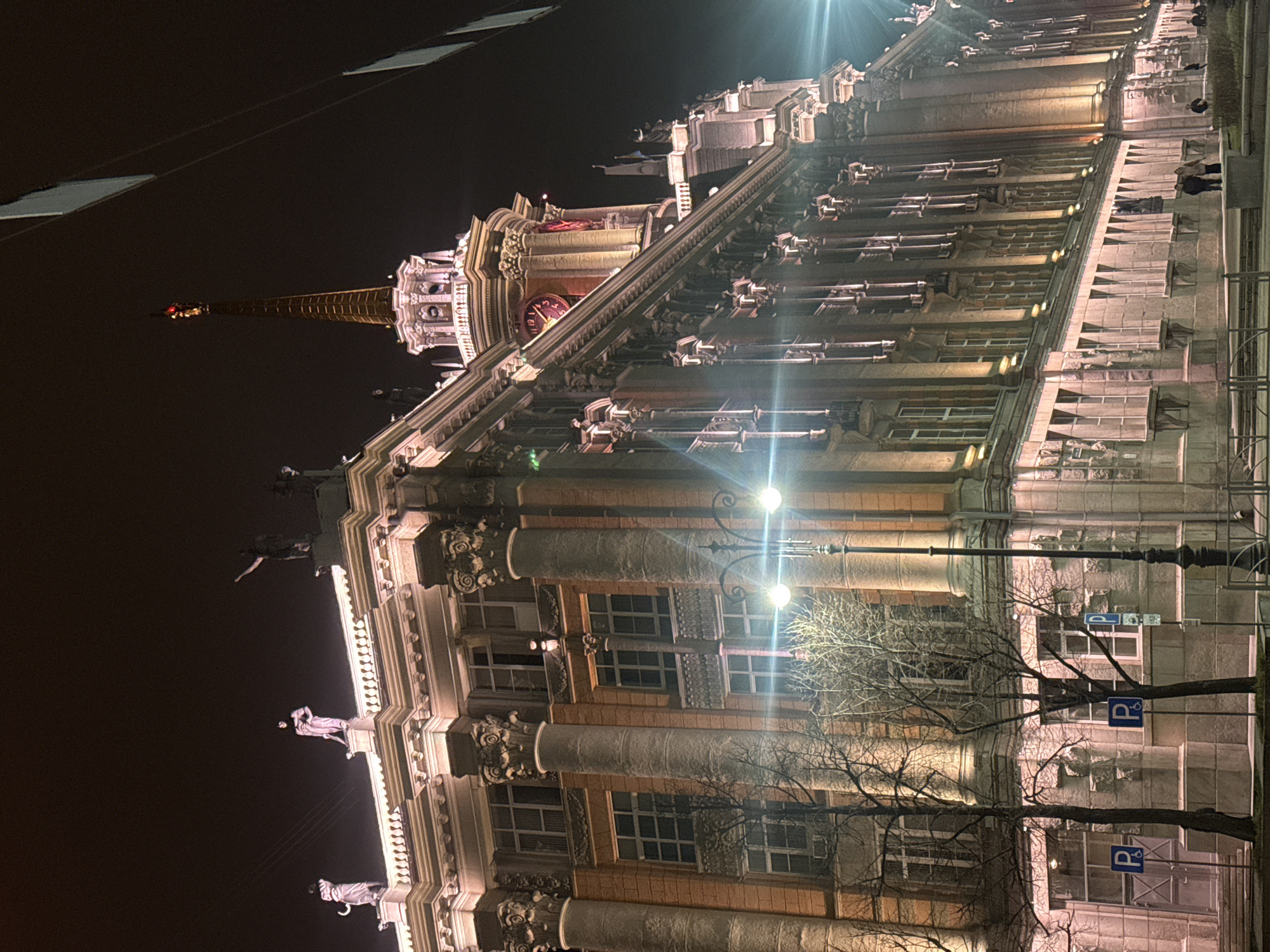This image depicts a grand, ornate building illuminated at night in an urban setting, contextually identified as Yekaterinburg, Russia. The multi-story structure is heavily detailed with classical architectural elements.
At the pinnacle of the building is a tall, slender spire, topped with a small, glowing beacon. Below the spire, a decorative tower section features a large, round clock face with Roman numerals, emitting a warm, reddish-orange light. The upper levels and roofline are adorned with numerous statues, including figures resembling gargoyles, and intricate carvings that catch the light.
The building's main facade showcases multiple rows of windows, many set within recessed architectural bays. Prominent fluted columns extend vertically across several stories, lending a stately and imposing character to the edifice. The facade appears to be constructed from a mix of light-colored stone or concrete and sections of reddish-brown material, all brightly lit by upward-pointing floodlights. A strong light source creates a noticeable lens flare in the scene.
At street level, a paved sidewalk is visible in the foreground. Several bare deciduous trees stand along the street, their branches silhouetted against the illuminated building and the night sky. Two streetlights with globe fixtures cast their own light. Visible on the sidewalk are blue parking signs, each displaying a white "P" for parking and a white wheelchair symbol, indicating accessible parking. A few indistinct figures can be seen on the sidewalk near the base of the building. The sky is dark, characteristic of nighttime, emphasizing the artificial illumination of the architecture.
