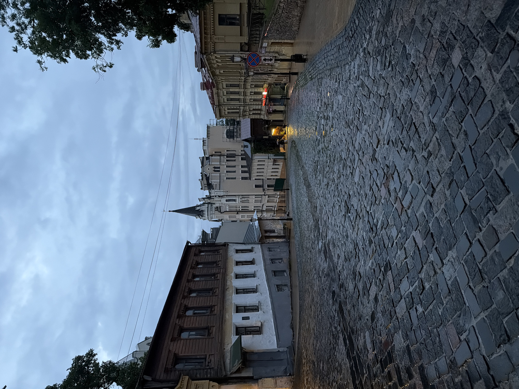 A wet cobblestone street slopes upwards between old buildings under a cloudy sky in Kyiv, Ukraine. A lone person holding a black umbrella walks away from the camera in the middle distance. The architecture suggests an older European city, with a mix of wooden and stucco facades. The street is slick with rain, reflecting the dim light from street lamps and windows.