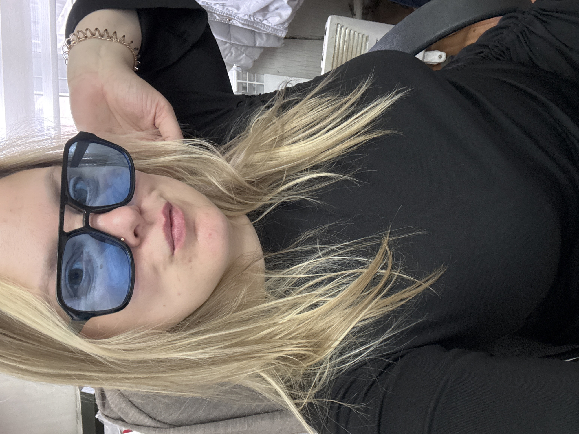 A fair-skinned woman with blonde hair and blue sunglasses is lying down, facing the camera. She wears a black top and a gold bracelet on her left wrist. A nose piercing is visible. Behind her are partially visible bedding, a white radiator, and a grey chair. The location is not identifiable as Noviy, Russia, based solely on the image.