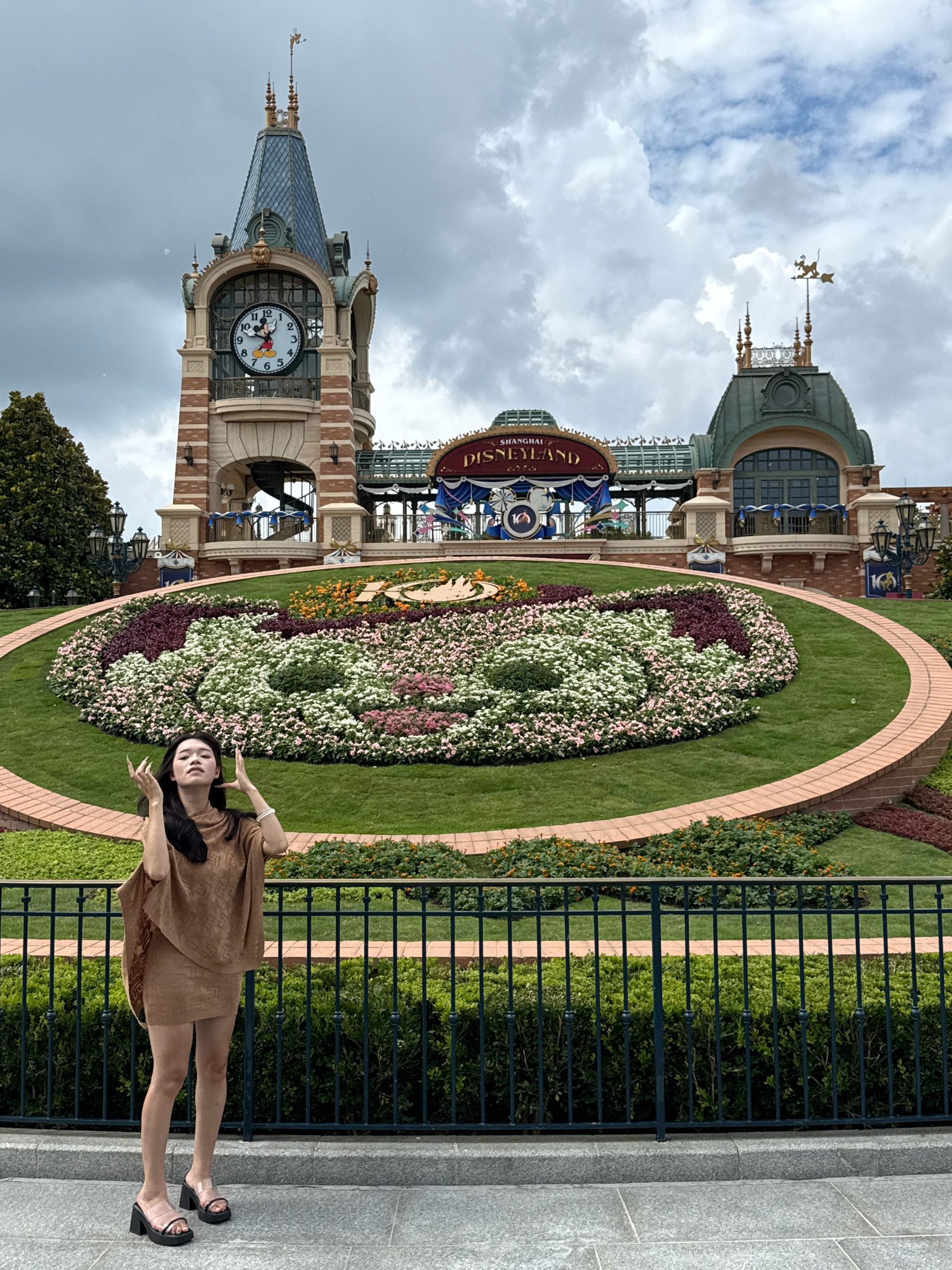 In the city of Pudong, China, outside Shanghai Disneyland, a young woman stands on a paved walkway behind a black metal fence. She is wearing a brown poncho and a matching short skirt, with black open-toe platform sandals. Her hands are raised and open, and her eyes are closed as she looks upwards. Behind her, a large, circular flowerbed displays a vibrant arrangement of pink, white, and dark red flowers against a backdrop of green grass. In the background, a clock tower with a Mickey Mouse face stands prominently, topped with a weather vane. The Shanghai Disneyland sign is visible on a structure behind the flowerbed. The sky is overcast with clouds.
