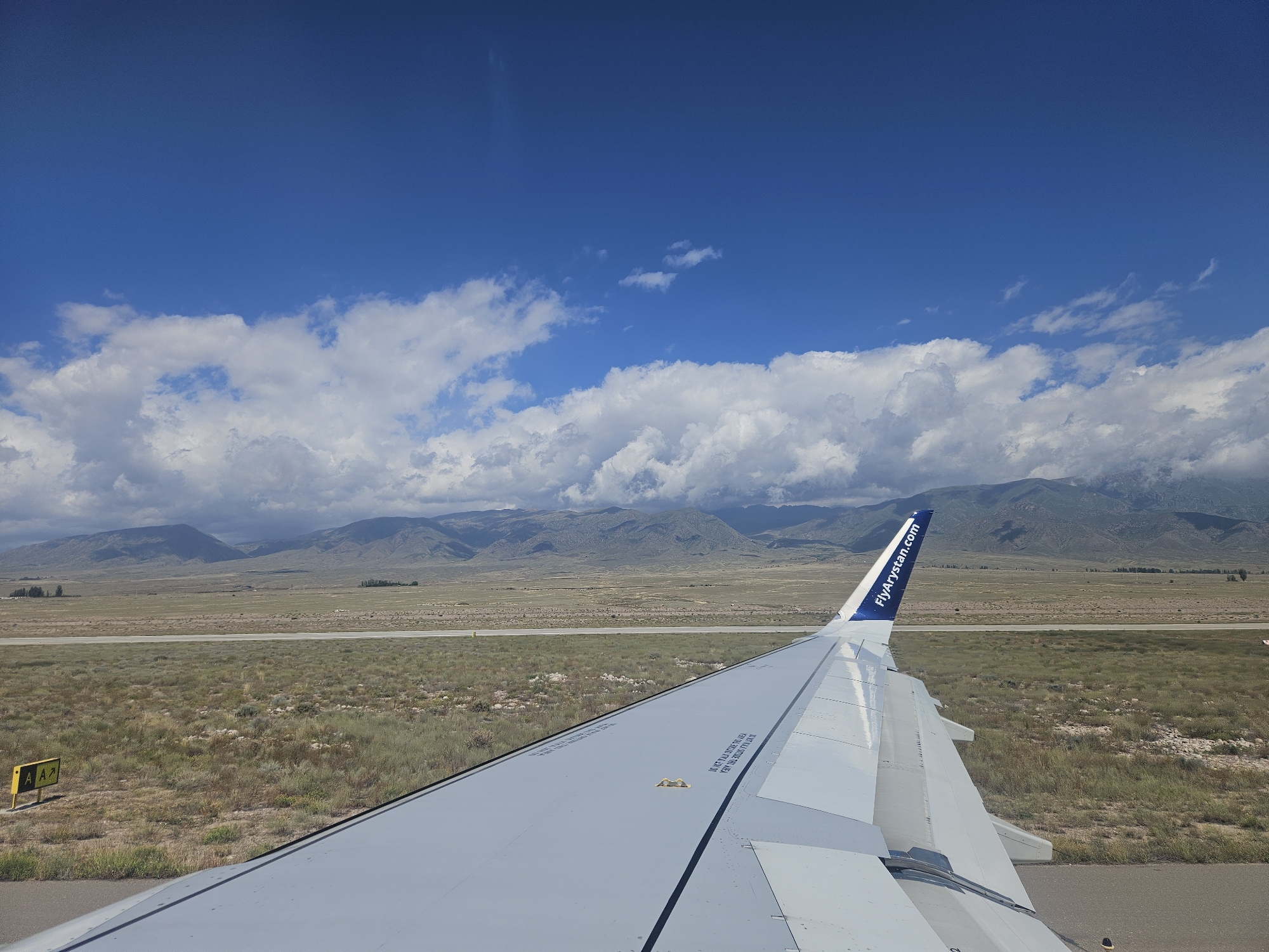 This is a perspective shot from inside an airplane looking out of a window at an airport in Tamchy, Kyrgyzstan. The airplane wing is visible in the foreground, taking up the lower right portion of the frame. The wing is light gray with dark blue accents and has the text "FlyArystan.com" on its tip. Beyond the wing, a runway stretches across a grassy and arid landscape. In the distance, mountains form a backdrop under a bright blue sky dotted with white clouds. The scene suggests the airplane is either preparing for takeoff or has just landed.