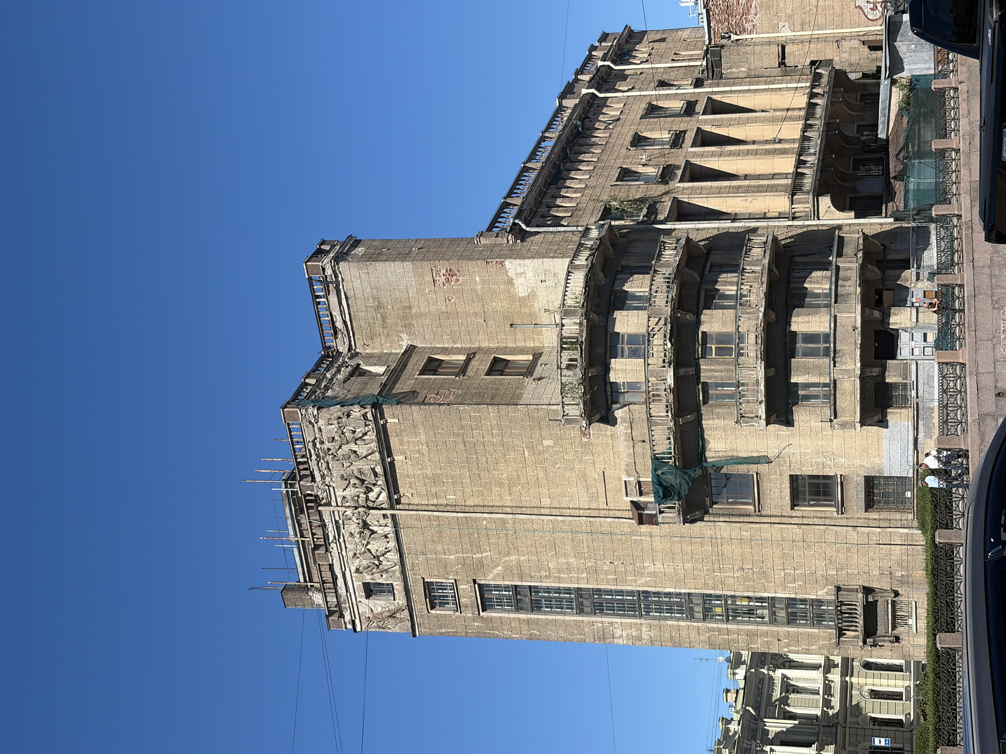 The image captures a view of the Singer House in Saint Petersburg, Russia, a prominent Art Nouveau building located at Nevsky Prospekt 28. The perspective is from across the Griboyedov Canal, looking up at the building's distinctive facade.
The Singer House is a multi-story structure characterized by its prominent glass dome, which is not visible in this particular shot. The building's exterior is constructed of stone, with a mix of architectural styles, including Art Nouveau elements and more classical features. The facade displays several floors with numerous windows, some of which are tall and narrow, suggesting an industrial or office purpose. There are also several balconies with decorative railings, some of which appear to be in disrepair or covered with green netting.
The upper part of the building features a decorative frieze with reliefs of human figures, suggesting a narrative or allegorical theme. The roofline is topped with a balustrade and various structures, possibly ventilation or antenna systems.
In the foreground, the Griboyedov Canal is partially visible, with a decorative metal fence along its embankment. Several people are seen walking along the embankment, appearing as small figures in the frame. The lower part of the image shows the roof of a black car.
The sky above is a clear, vibrant blue, indicating a sunny day. The overall impression is of a historic urban environment, with the Singer House standing as a significant landmark against the clear sky. The building exhibits signs of age and weathering, but its architectural grandeur remains evident.