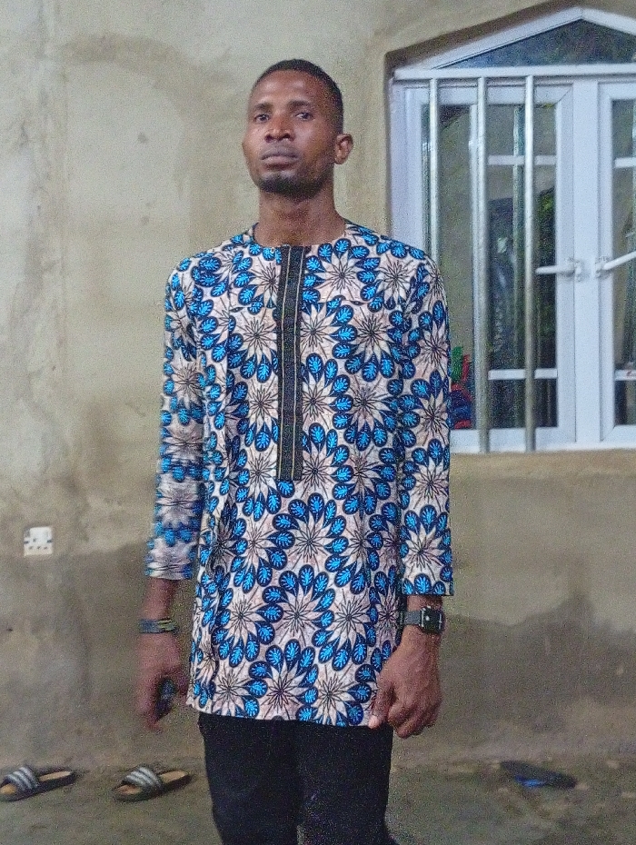 An adult man stands centrally, facing the camera with a neutral expression in what appears to be an indoor or covered outdoor area in Nando-Ikem, Nigeria. He has short dark hair and a neatly trimmed beard or stubble.
He is wearing a long-sleeved tunic shirt adorned with a vibrant, intricate pattern of repeating blue and light-colored floral or paisley-like motifs against a cream or beige background. A prominent black vertical strip with decorative trim runs down the center of his chest. He pairs the tunic with dark trousers or jeans. On his left wrist (viewer's right), he wears a dark-colored watch, and on his right wrist (viewer's left), a dark bracelet is visible.
The background features a plain, light-colored wall, possibly concrete or stucco. To the man's right, a white-framed window with vertical security bars is visible. Behind his left shoulder, a light switch or electrical outlet is mounted on the wall. The ground beneath him is also light-colored.
On the ground to the man's left, two pairs of slide sandals are visible. The sandals are dark with the distinctive three white stripes of the Adidas brand. The lighting in the image suggests it might be an indoor setting or during the evening hours. The man appears to be posing for a photograph.