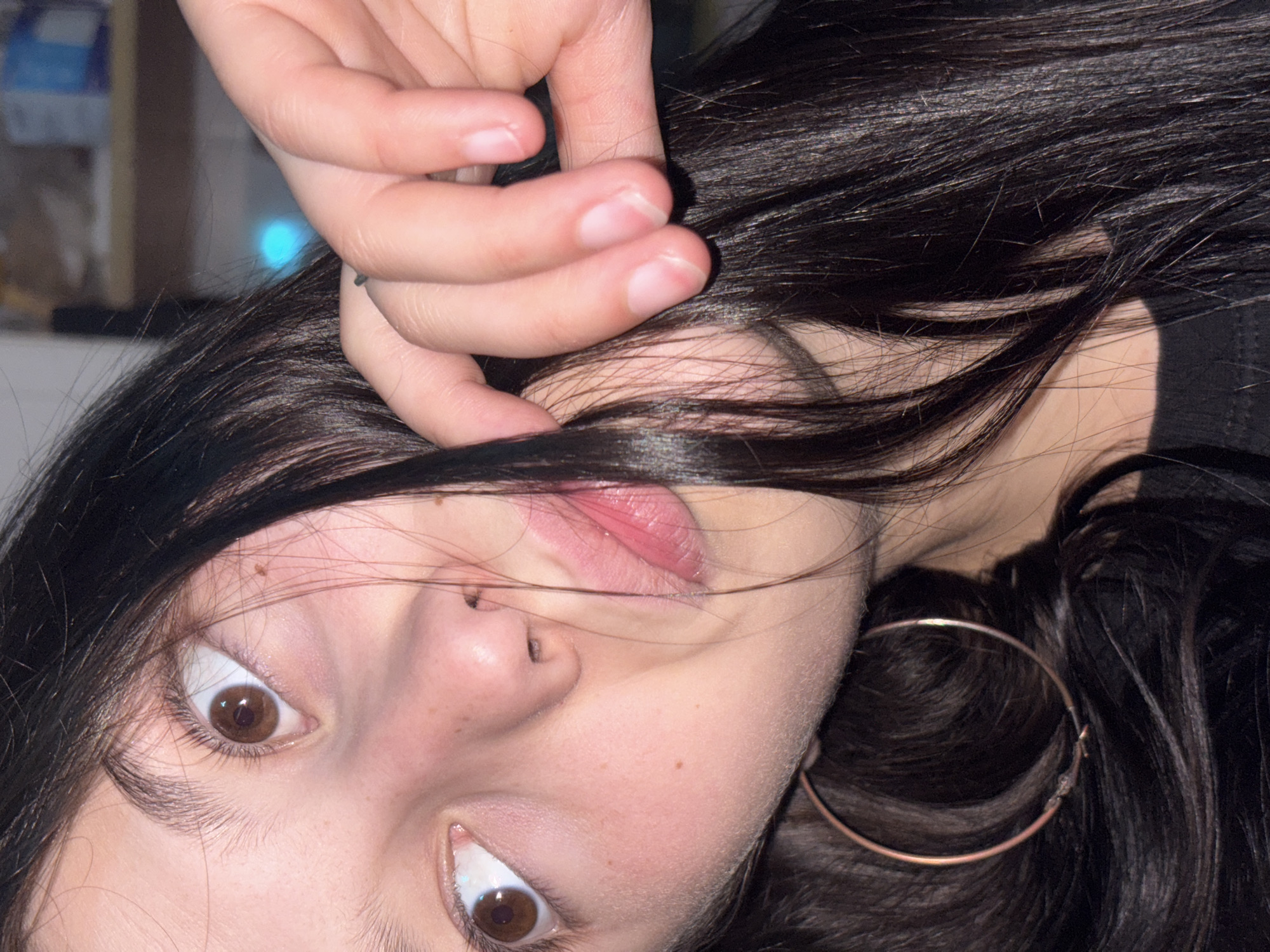 The image is a close-up, low-angle shot of a young person's face, with their head tilted to the side. Their eyes are wide open and looking upwards, with the left eye more prominently visible. They have dark hair that partially covers their face. A hand is reaching up towards their hair, with fingers slightly curled. One large hoop earring is visible on the lower left side of the image. The lighting is warm and suggests an indoor setting, possibly with artificial light sources. There are some subtle skin blemishes and freckles visible. No specific location in the city of Fokino, Russia can be determined from the visual information provided.