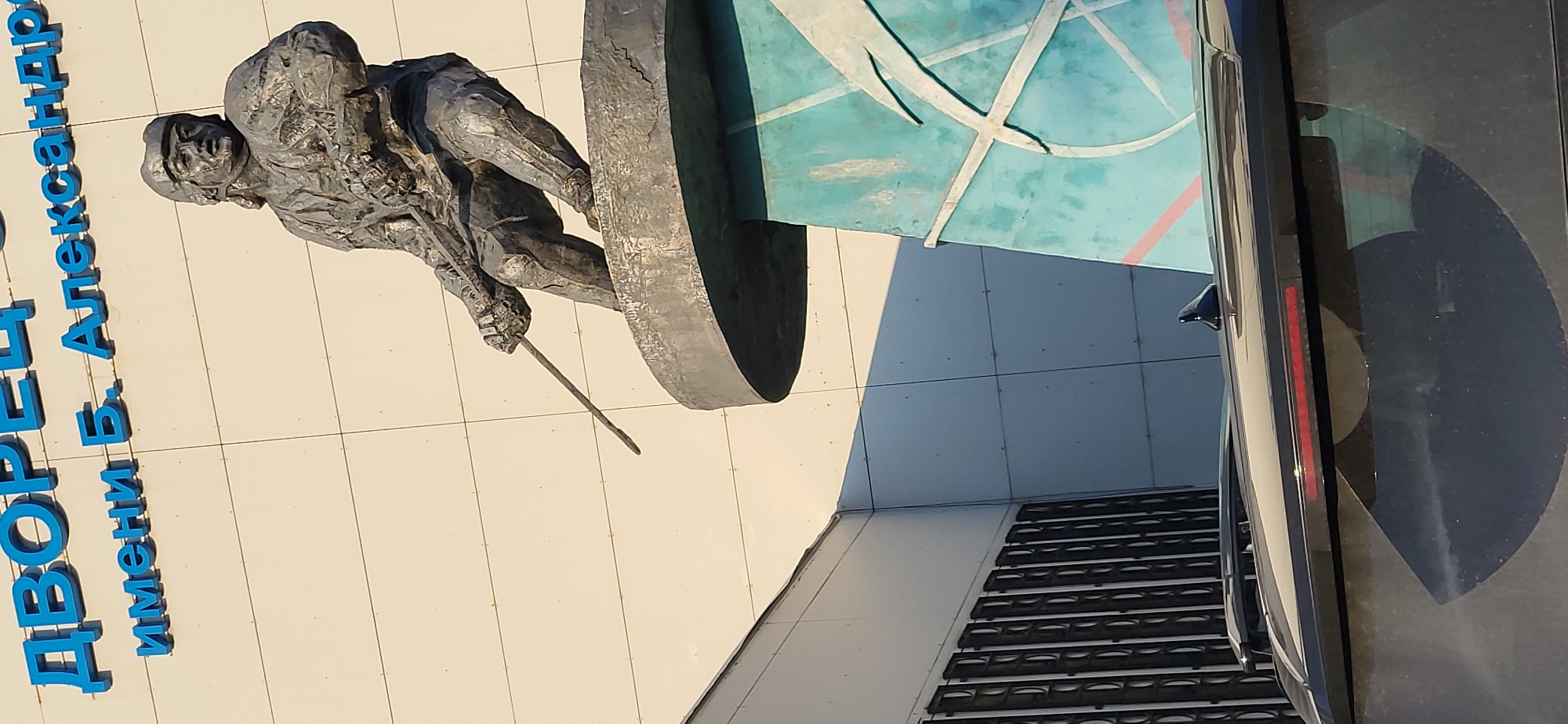 The image shows a statue of a person in a dynamic pose, holding a long, thin object, likely a sword or a lance, in their right hand. The statue is made of metal and is positioned against a modern building with blue lettering that reads "Дворец имени Б. Алексеандра". The building's facade is made of cream-colored panels. Below the statue, there is a teal-colored structure with white and pink lines forming a geometric pattern. In the foreground, the top of a dark-colored vehicle is visible, with its windshield reflecting the sky and the building. The scene appears to be outdoors, likely in a public space within the city of Oskemen, Kazakhstan, given the text on the building. The lighting suggests it is daytime, with sunlight casting shadows.