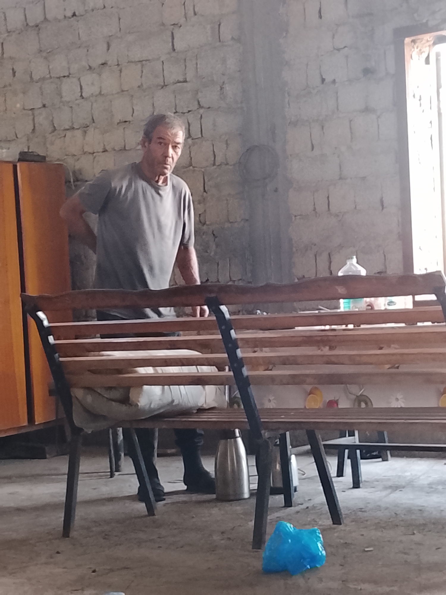 A middle-aged man with graying hair and a concerned expression is standing in a dimly lit, unfinished room with brick walls. He is wearing a grey t-shirt and dark pants, with his right hand on his lower back. In front of him is a wooden bench with black metal legs, behind which is a table covered with a patterned cloth. A large silver thermos and a blue plastic bag are visible on the floor. The room appears to be in a state of disrepair or under construction. The image captures a moment of pause for the man, who seems to be experiencing discomfort or pain.