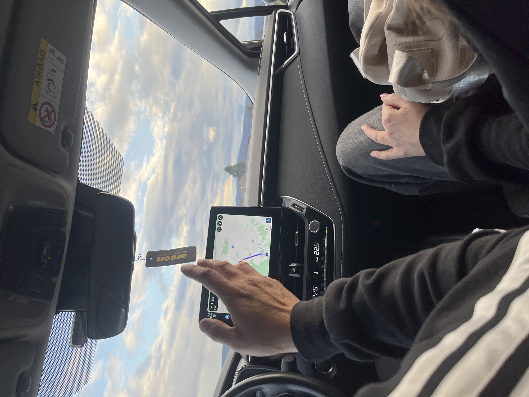 The image depicts the interior of a car, specifically the dashboard area. A man is touching the screen of a GPS navigation system, which displays a route. The display is showing a location in or near Staro Selo, Bulgaria. An air freshener is hanging near the rearview mirror. The driver is wearing a black hoodie with white stripes. A passenger is visible in the top right of the image, seated. The sky is visible through the windshield. The vehicle's air vents and temperature control panel are visible on the dashboard.