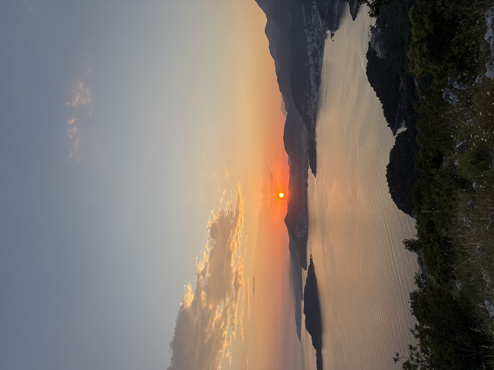 The image captures a sunset over a body of water near mountains. The sun is a bright orange orb, partially obscured by a distant mountain range. The sky fades from blue to orange near the horizon, with some clouds present. The water reflects the sunset colors. The right side of the image shows trees and vegetation on a mountain slope. The location appears to be in or near the city of Boreti, Montenegro, based on the surrounding landscape and the orientation of the water and mountains. There are no people visible in the image. The overall scene is a natural landscape at sunset.